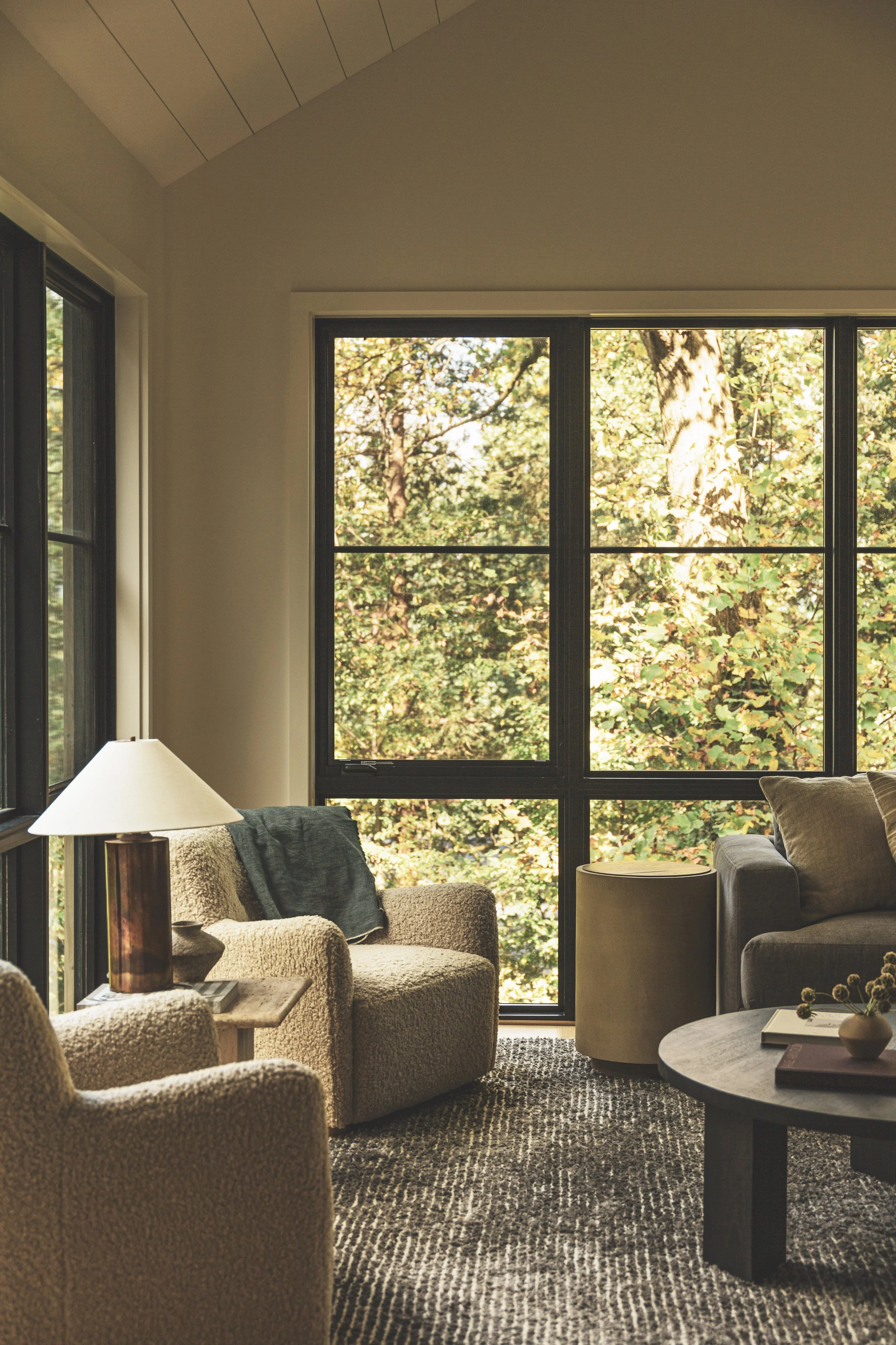 Living room with beige armchairs, a gray sofa, a round coffee table, a side table with a lamp, and large windows showing a view of trees outside.