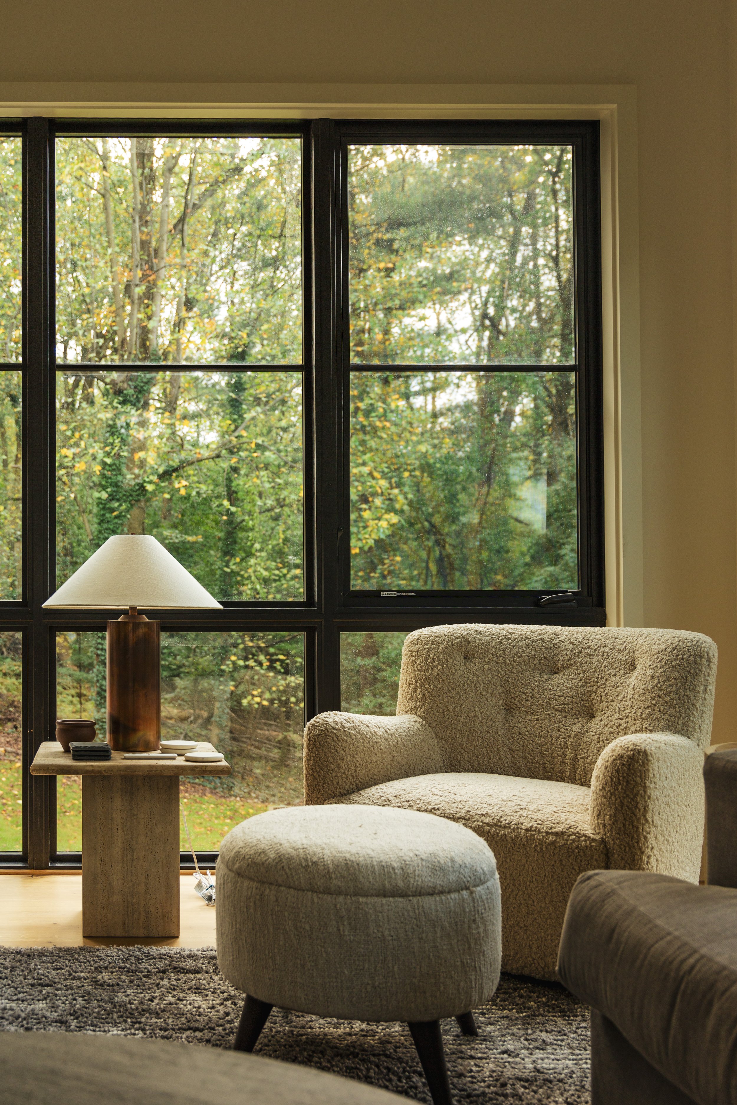 Living room with beige armchair, ottoman, side table with lamp, and large windows showing trees outside.
