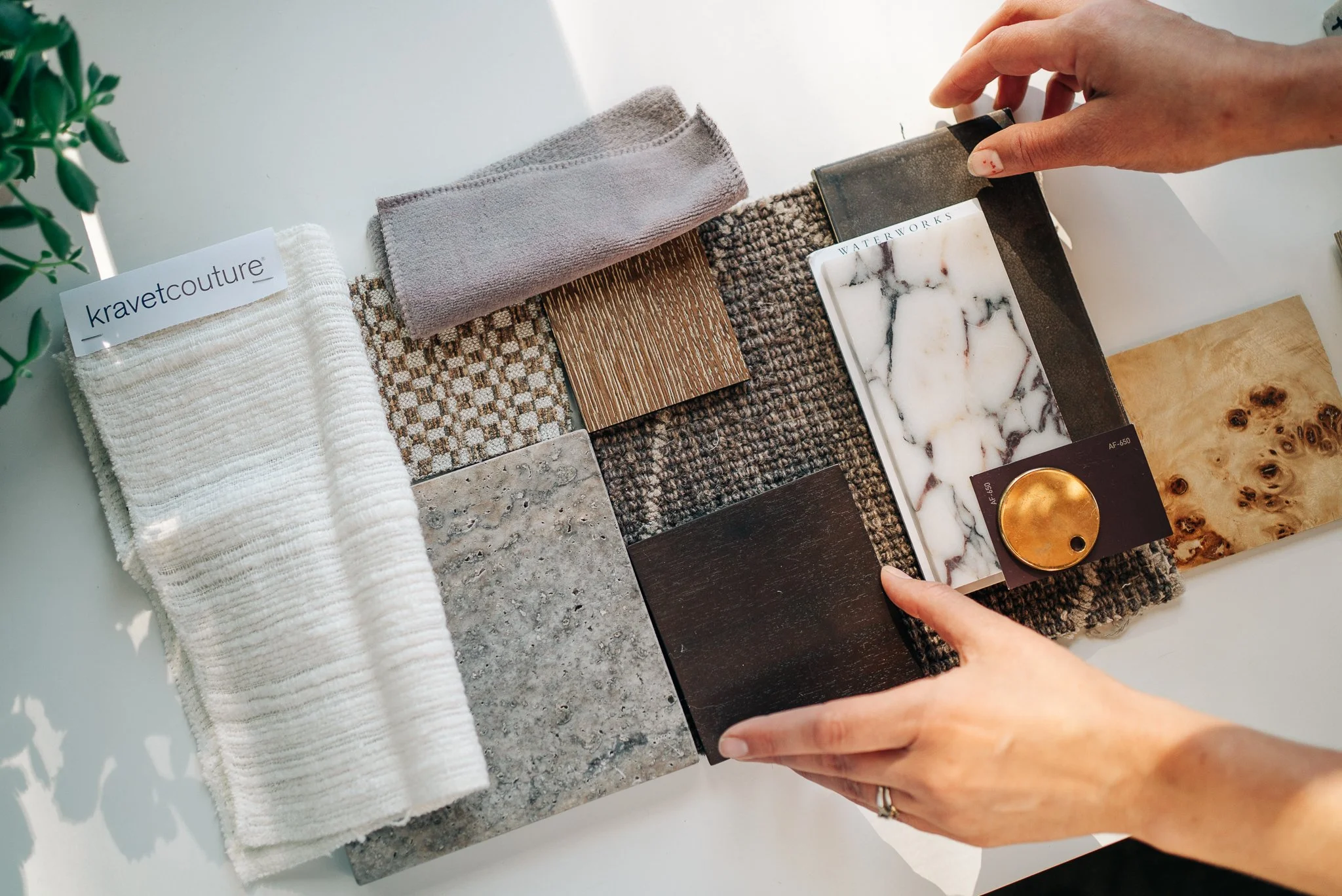 A person selecting a tile sample from a collection of various tile and fabric swatches on a white surface, including marble, wood, and fabric textures.