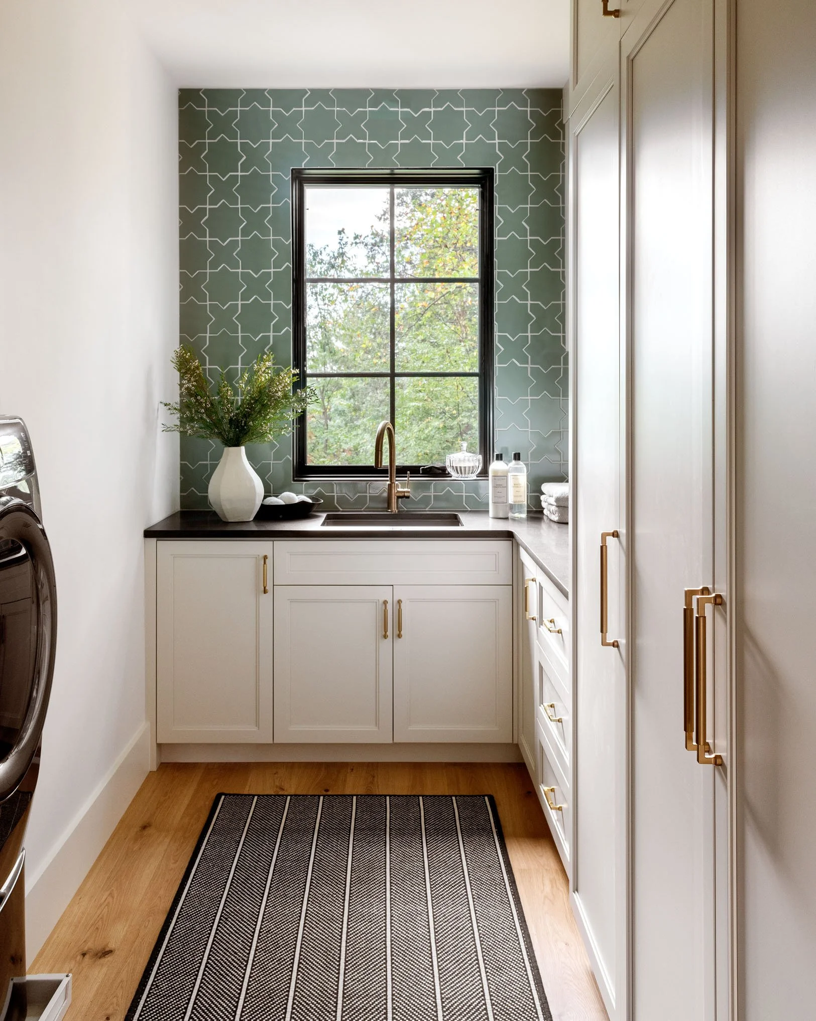 A laundry room with a window, white cabinets with gold handles, a rug on a wooden floor, and a countertop with toiletries and towels.