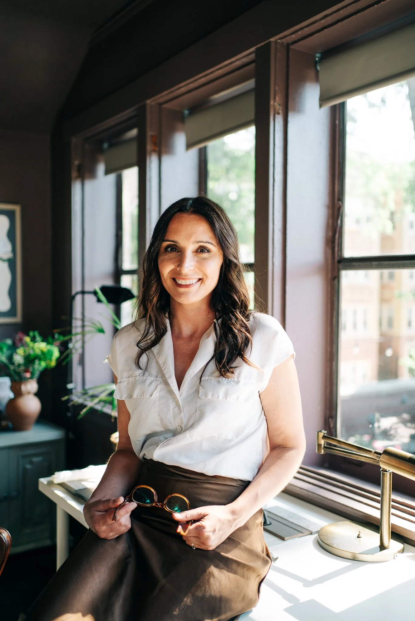 A woman with long dark hair, smiling, standing near a window with sunlight streaming in. She is wearing a white blouse and holding sunglasses in her right hand. The background includes a plant and a framed artwork.