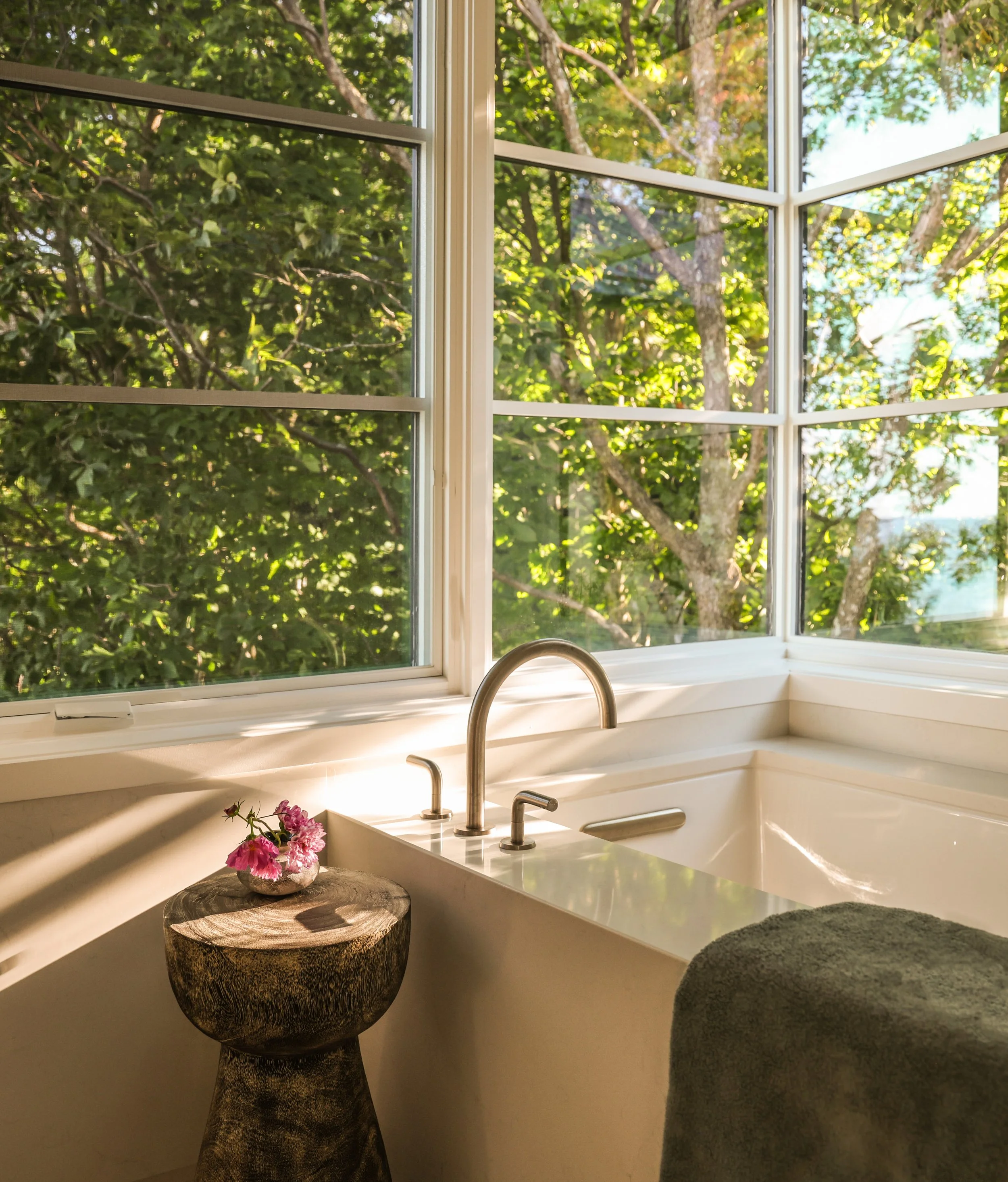 Inside a kitchen with a large window showing green trees outside, a white countertop with a silver faucet, a wooden stump side table with pink flowers, and a gray seat or cushion on the right.