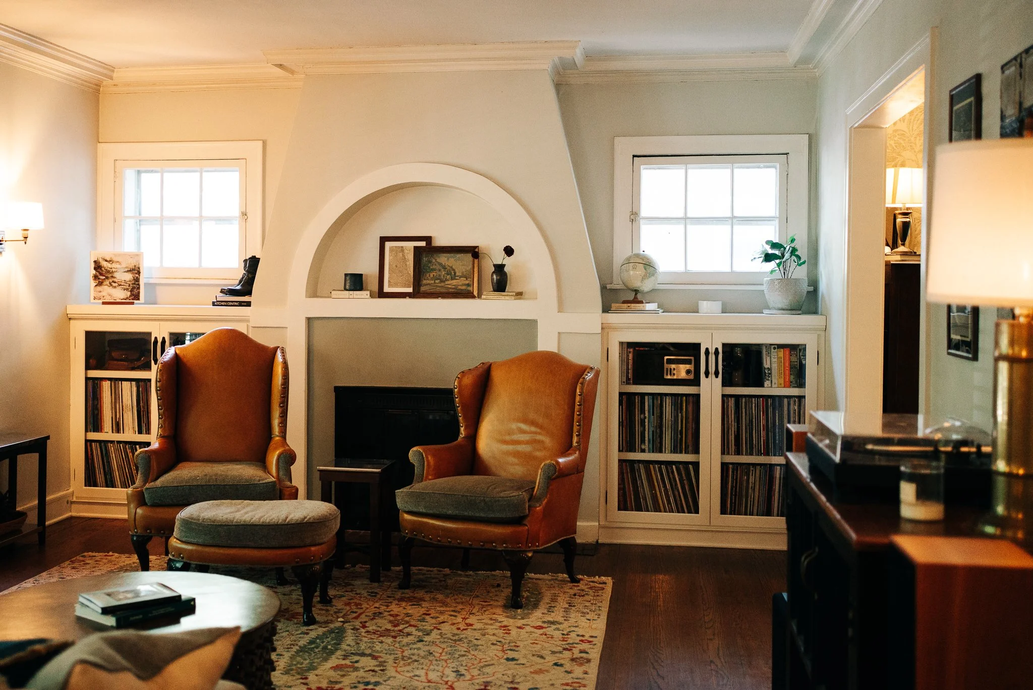 Living room with two armchairs, a footstool, bookshelves, windows, and various decorative items.