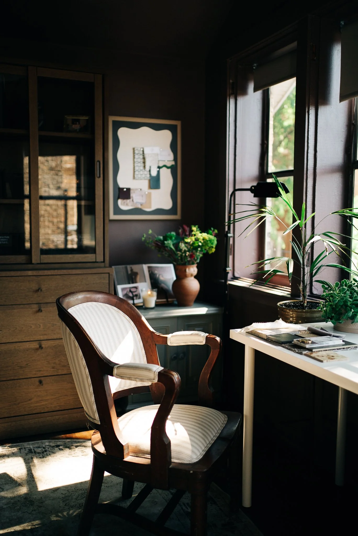 Cozy interior with a wooden cabinet, a framed bulletin board, a window with sunlight, a potted plant, and a table with photos, a candle, and books.