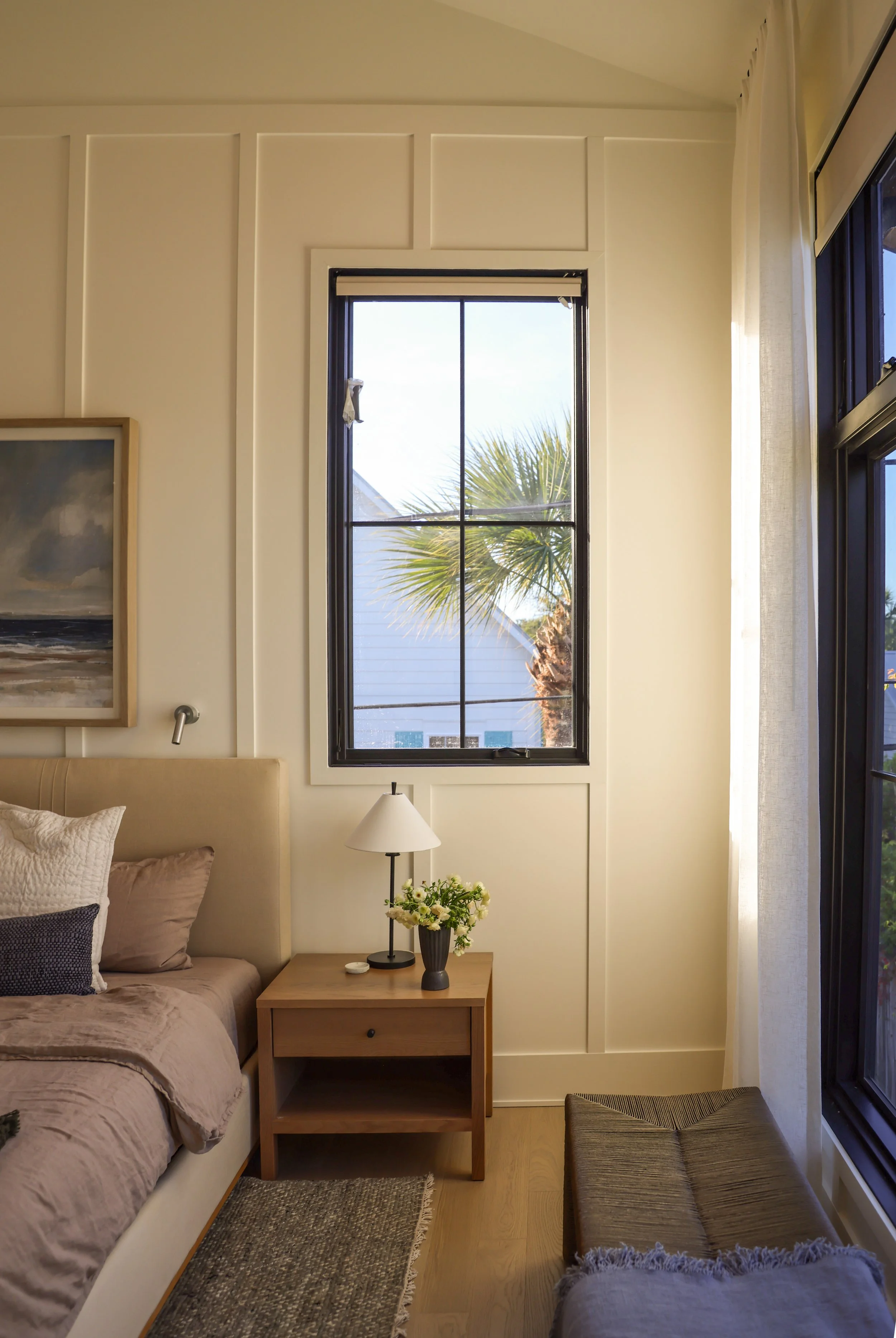 A cozy bedroom corner features a beige bed with pillows, a wooden nightstand with a white lamp and vase of flowers, a window showing a palm tree outside, and a window with a curtain on the right. There's a framed artwork on the wall and a small woven rug on the wooden floor.