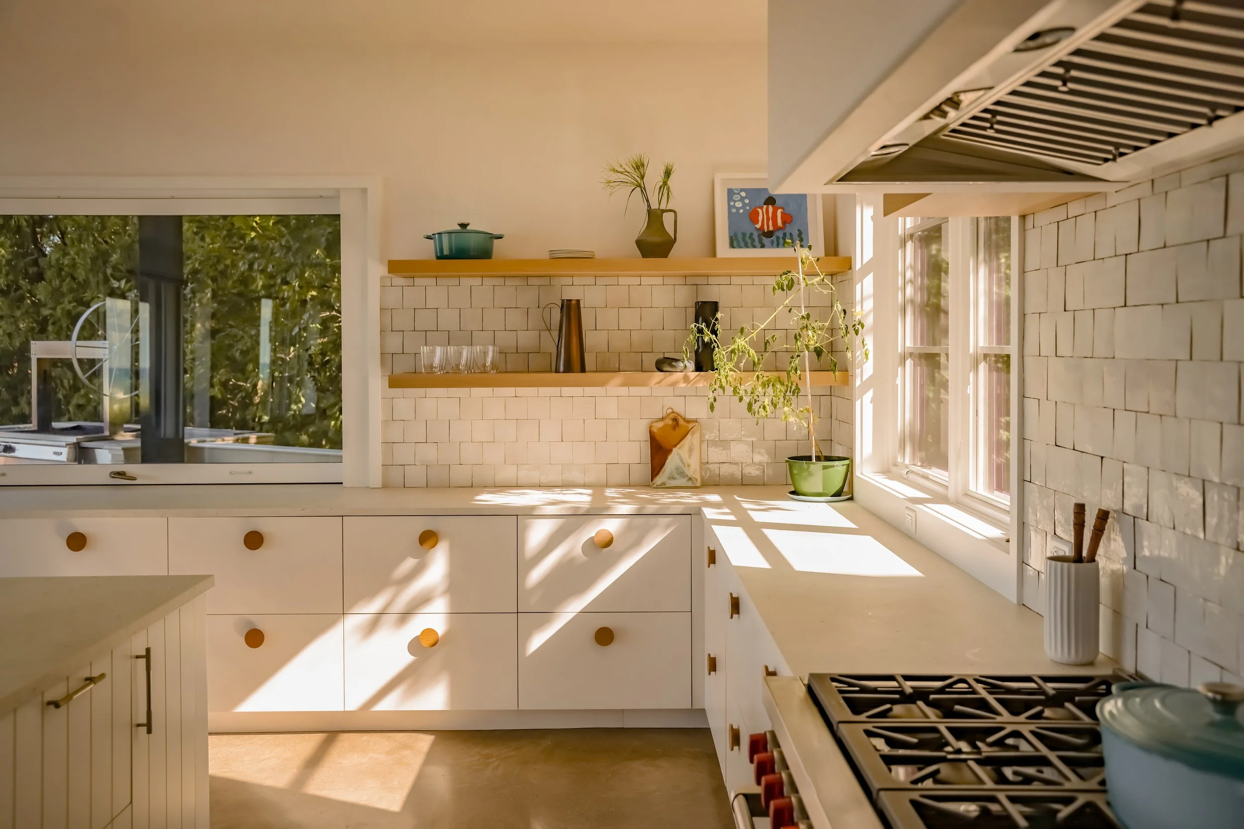 Sunlight streaming into a modern kitchen with white cabinets, a brick backsplash, open wooden shelves with kitchenware, a potted plant, and a window showing green foliage outside.