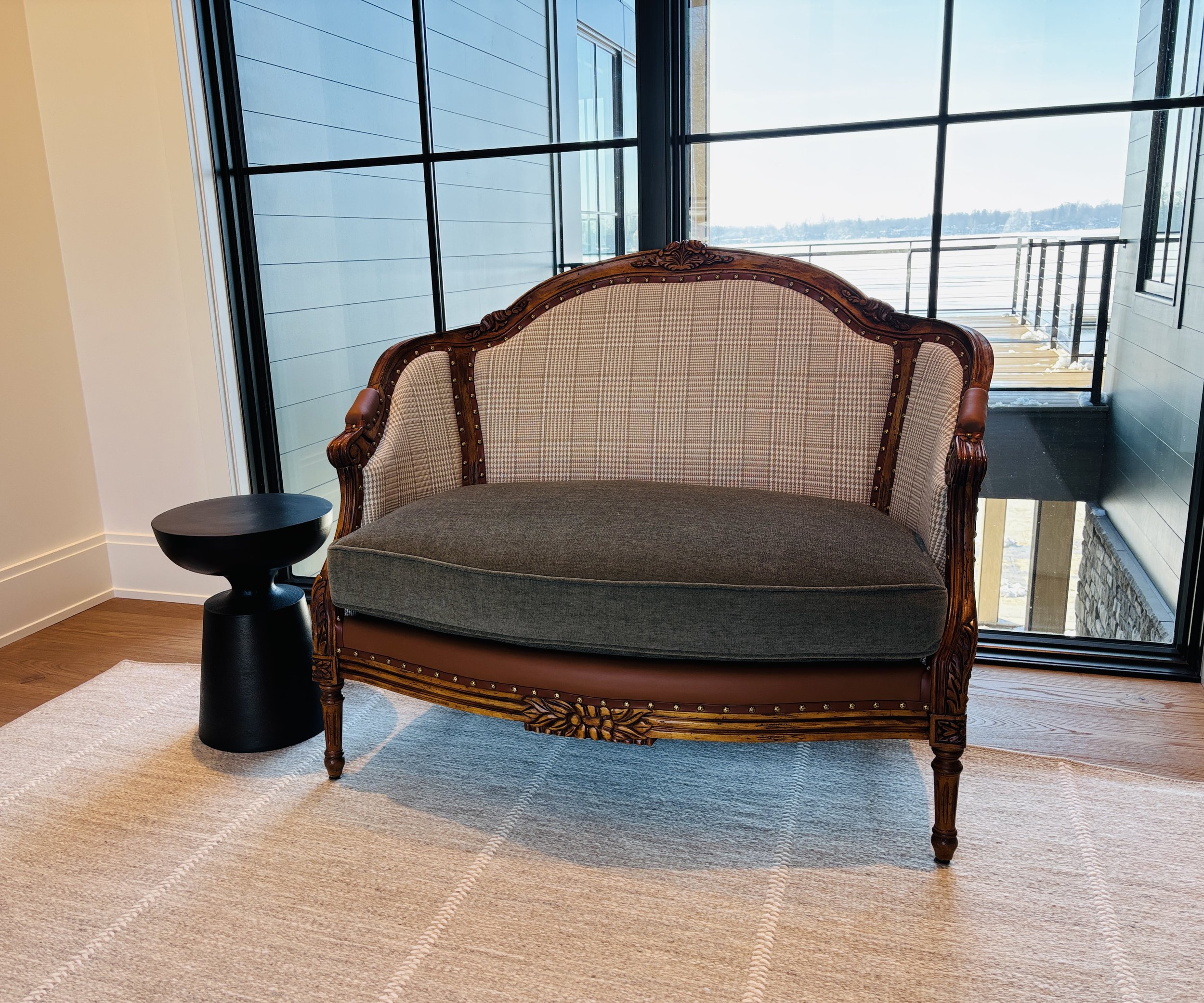 A vintage wooden settee with a cushioned seat and backrest, upholstering in beige patterned fabric, situated next to a small round black side table, in front of a large window with glass walls and a balcony showing a snowy landscape outside.