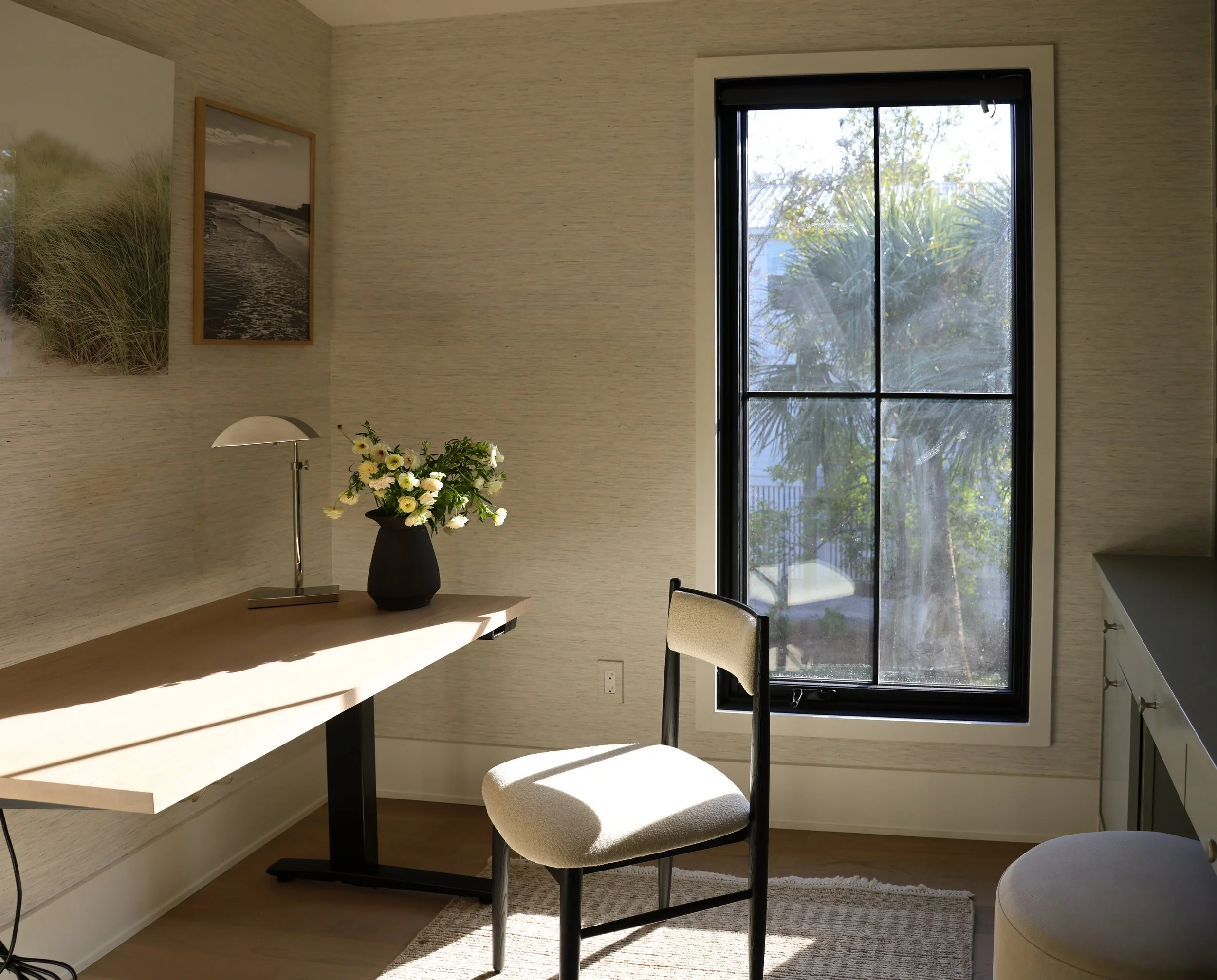 A cozy dining nook with a small table and upholstered chairs, featuring a large window with black framing that looks out onto lush greenery. The wall behind has two framed black-and-white landscape photographs, and there is a tall vase with white flowers on the table. A modern desk lamp is also on the table, with sunlight illuminating the space.
