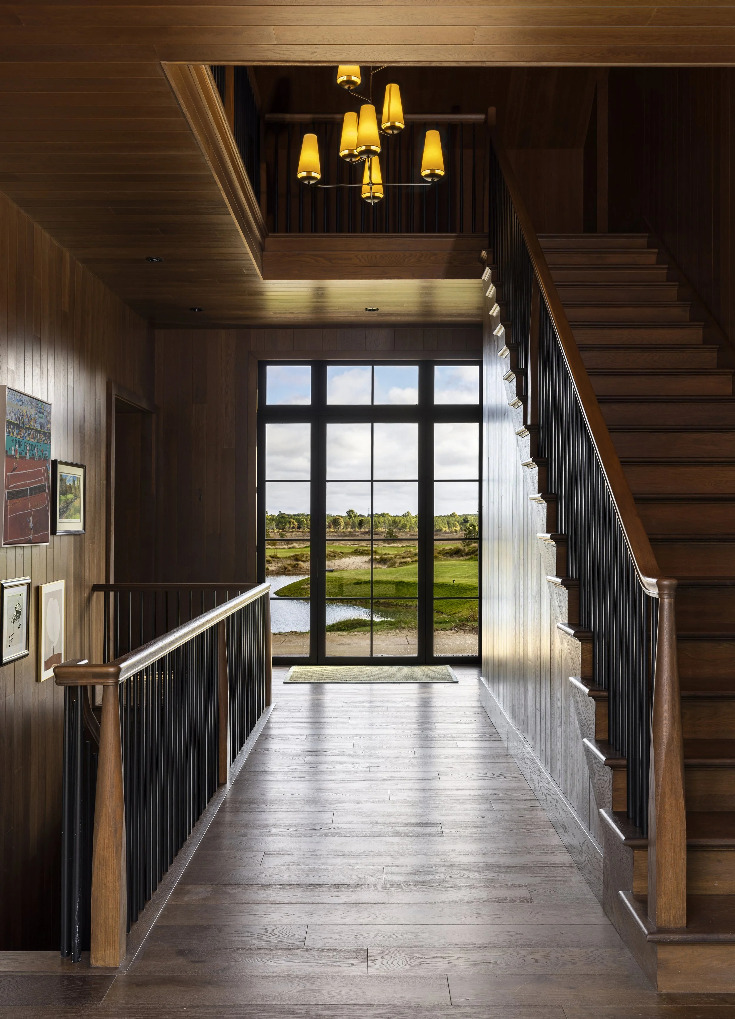 Image of a wooden-floored indoor space with a staircase on the right and a large window at the end showing a scenic outdoor view of a golf course with green grass, trees, and a partly cloudy sky.