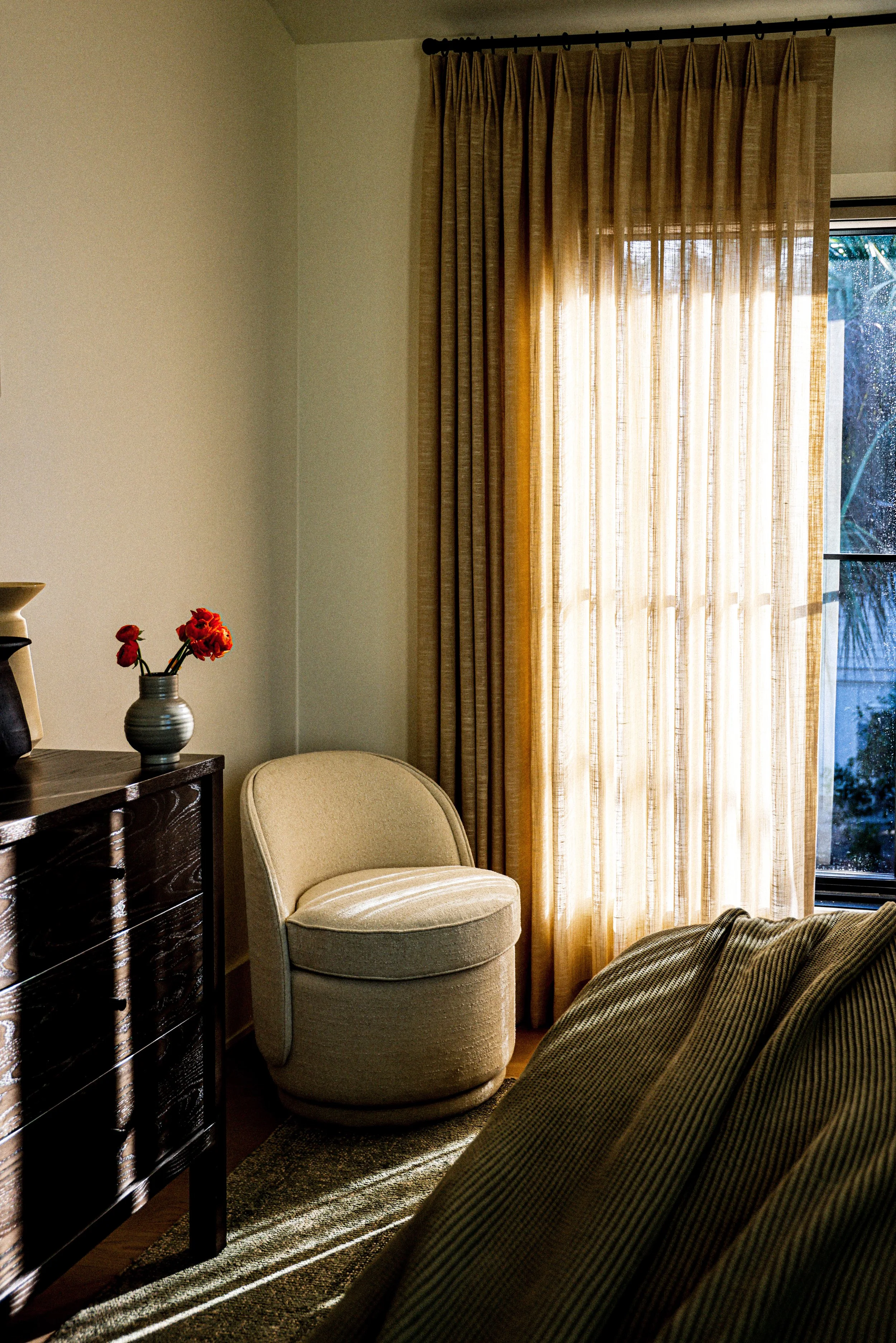Living room with beige armchair, dark wood dresser, beige curtains, and a window with rain outside.