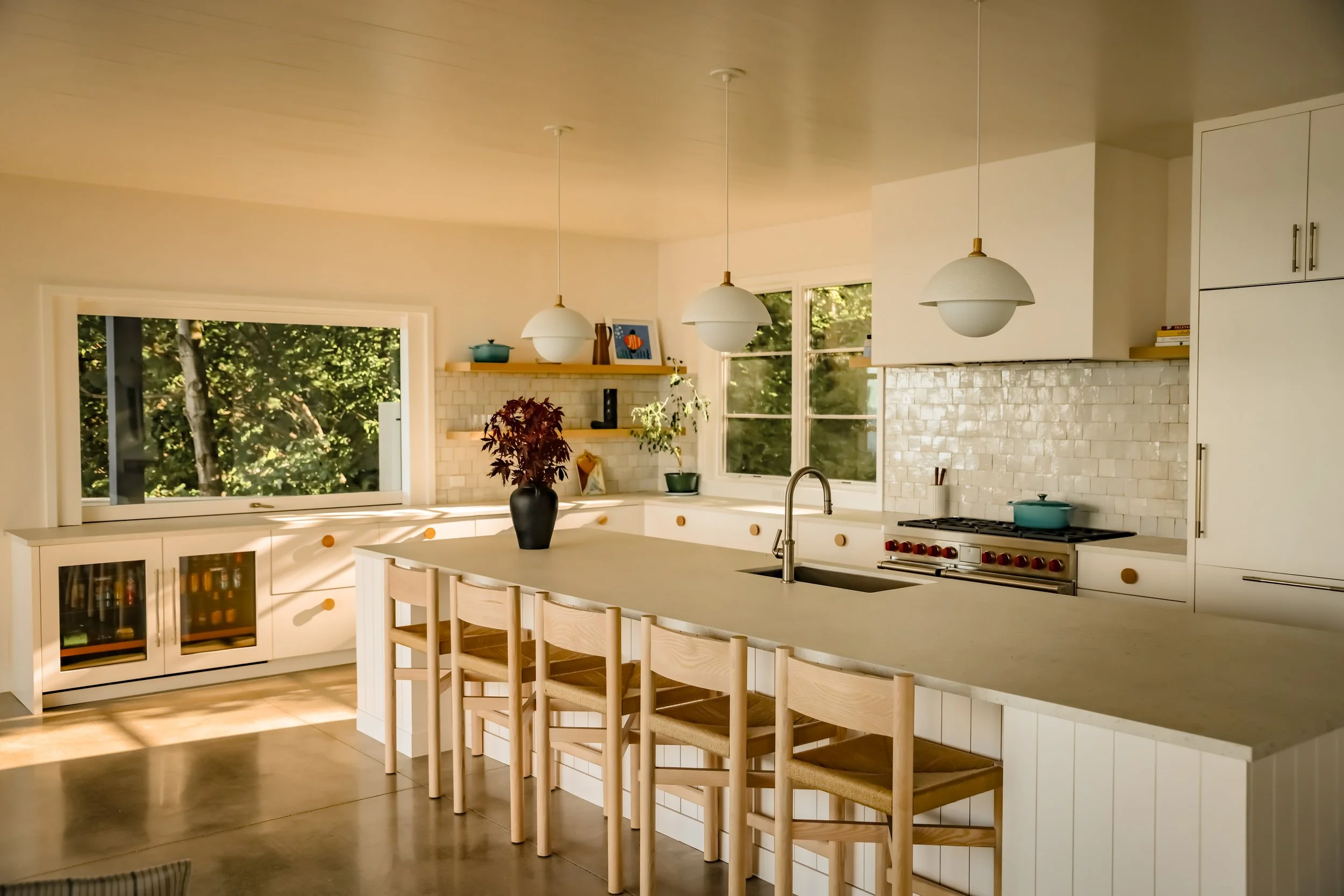 A modern kitchen with a large island, white cabinetry, pendant lighting, and large windows with a view of greenery outside.