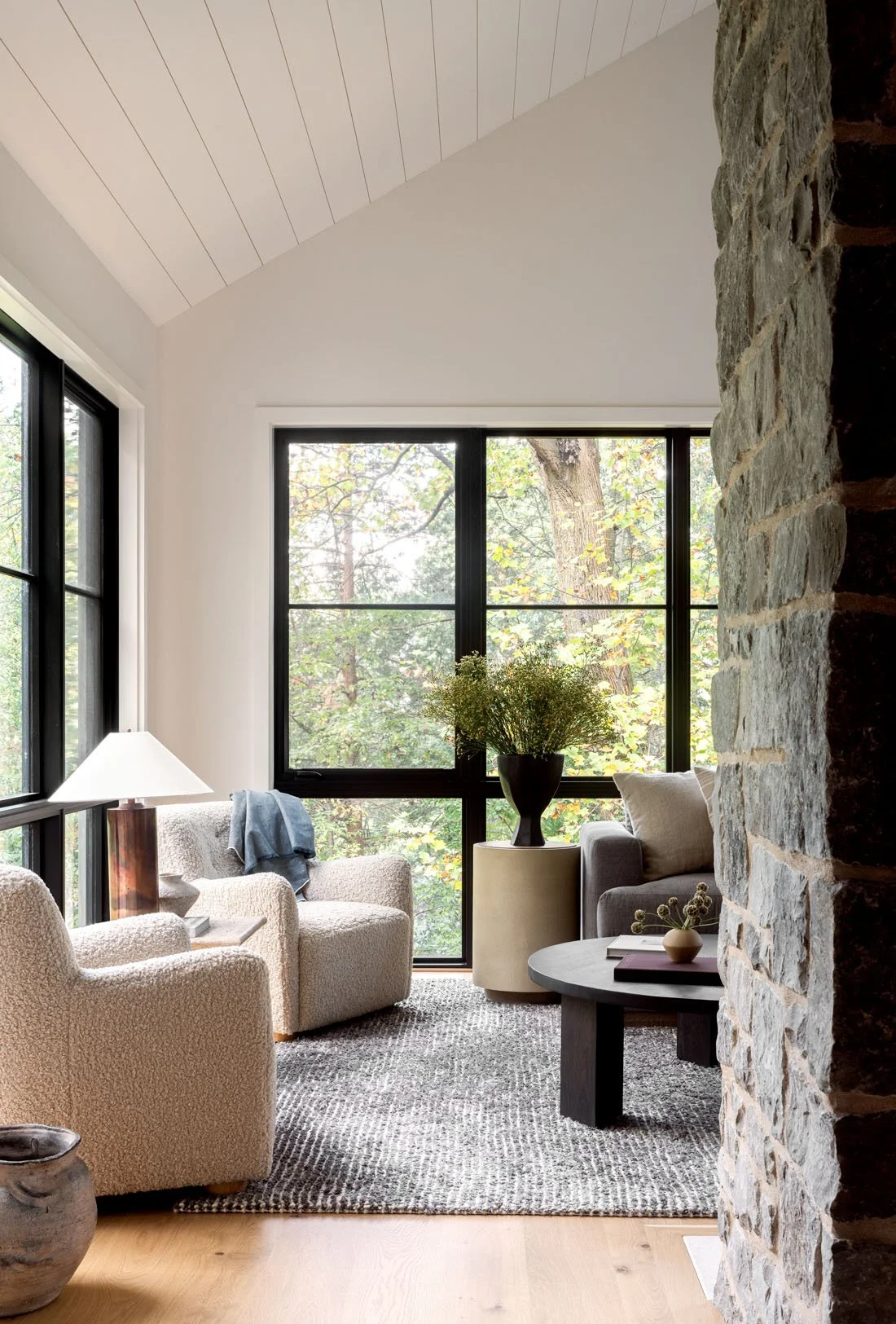 Living room with large windows showing trees outside, neutral armchairs, a side table with a lamp, a black vase with greenery, a round coffee table with decorative items, and a stone wall.