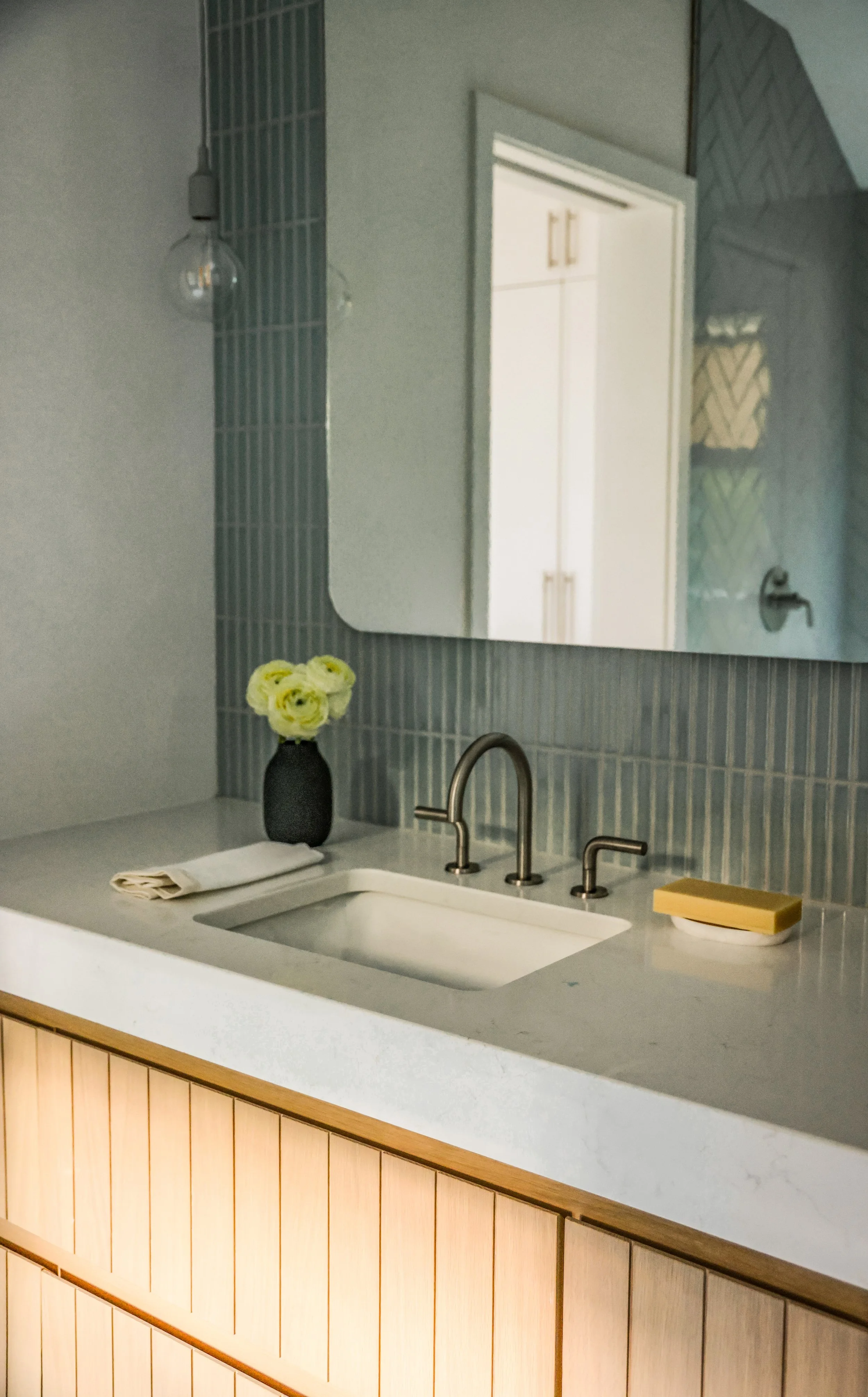 A bathroom sink with a faucet, a vase of yellow flowers, a folded towel, and a bar of soap on the white countertop. A mirror above the sink reflects a white door and cabinetry. The vanity has a wooden panel base, and the wall behind the sink is tiled with small vertical blue tiles.