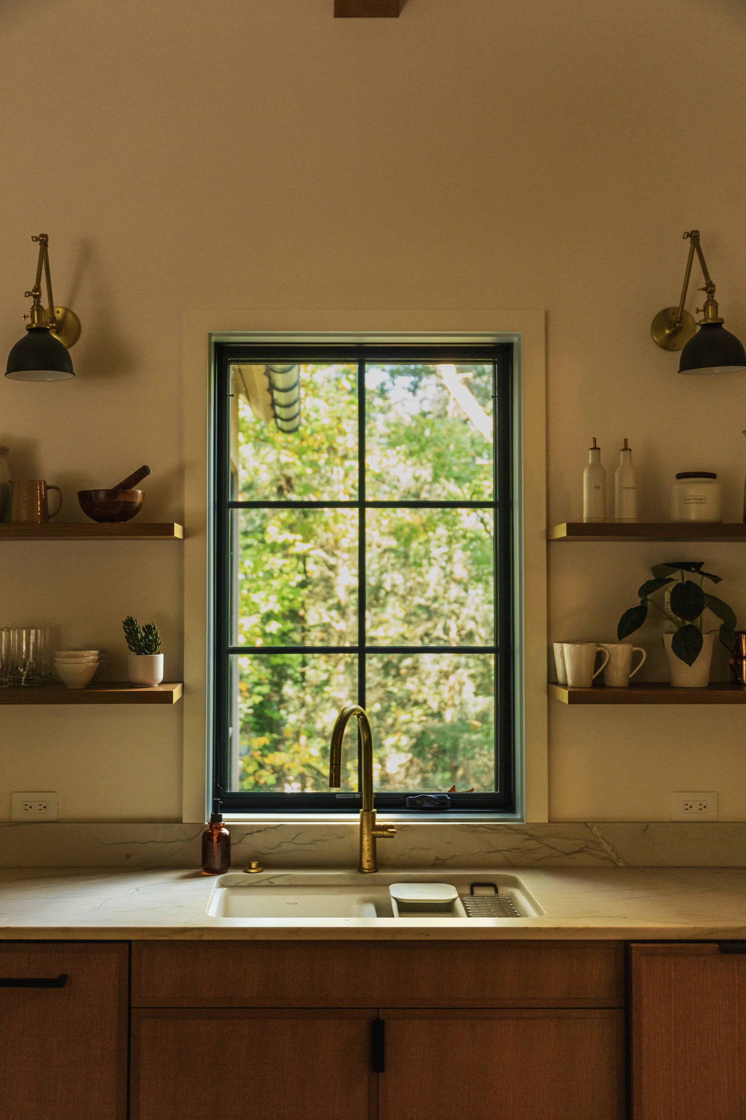 A kitchen window with a view of green trees outside, a gold faucet at the sink, and shelves with kitchen items like bottles, bowls, and potted plants, illuminated by two black and brass wall lamps.