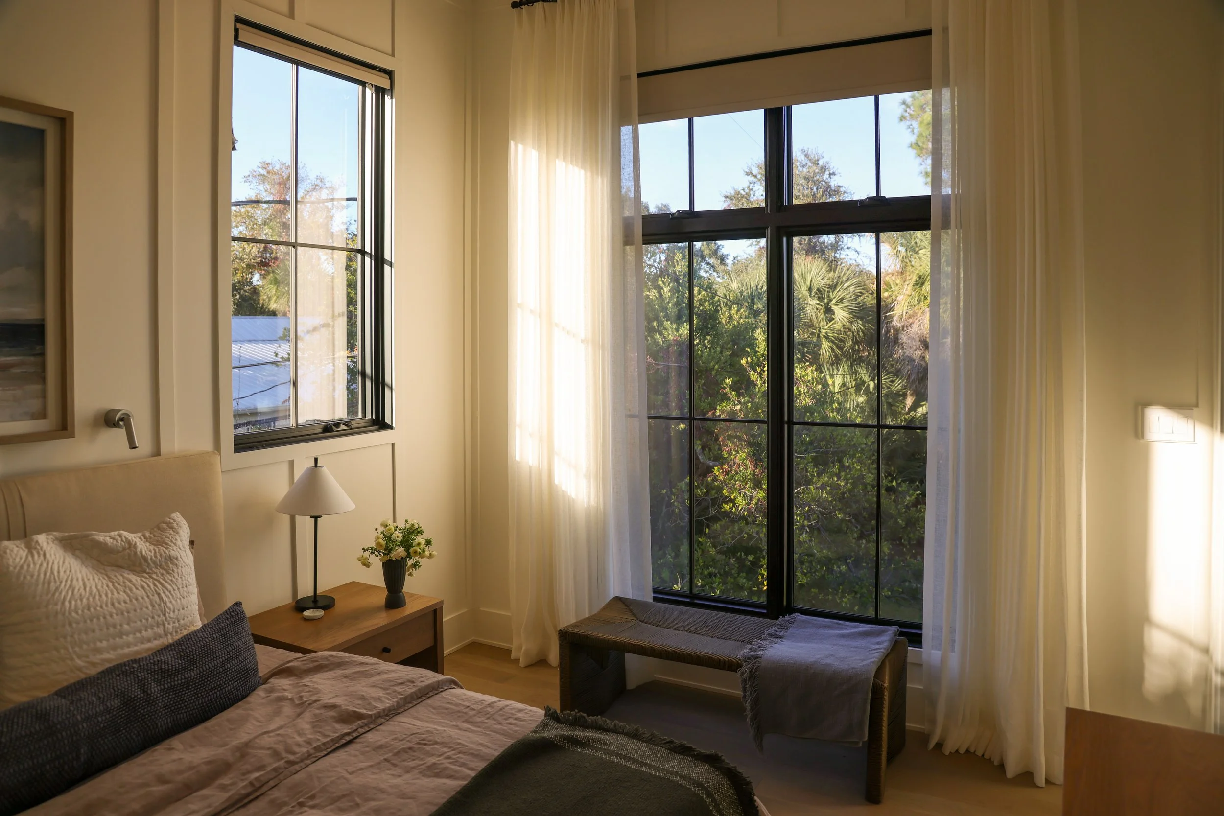 A cozy bedroom with two large windows letting in natural light, sheer white curtains, a bed with beige bedding and pillows, a wooden bedside table with a small flower arrangement, a table lamp, a small bench beneath the windows, and a light switch on the wall.
