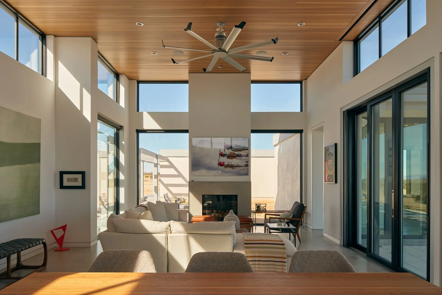 Modern living room with large windows, a ceiling fan, a fire place, a white sofa, and artwork on the walls.