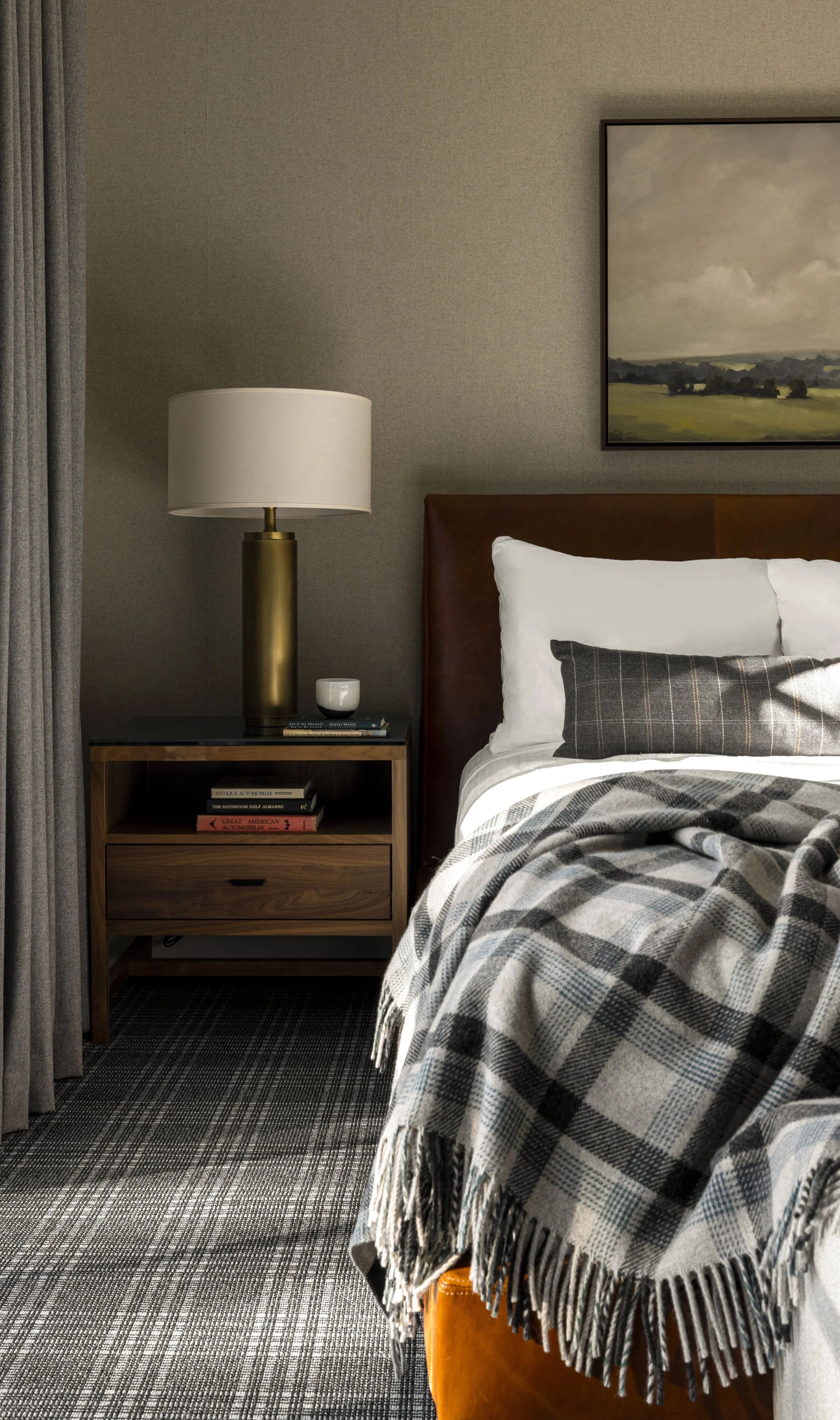 A bedroom corner with a nightstand, table lamp, stack of books, framed landscape painting, bed with pillows, and a plaid blanket.
