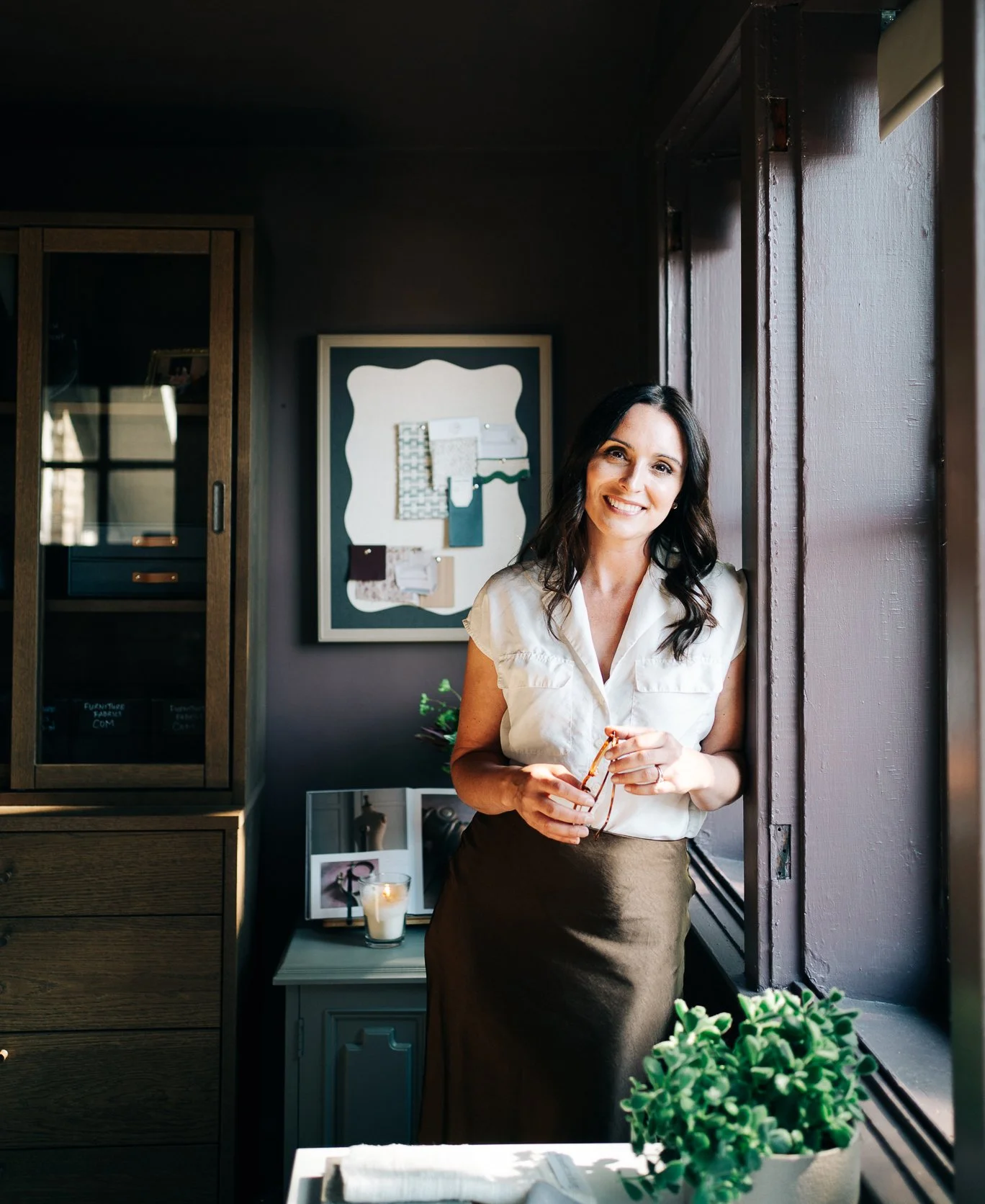 A woman standing by a window in a cozy room, holding glasses and smiling at the camera, with a bulletin board and framed photos on the wall behind her and a small table with a candle and a plant in the foreground.