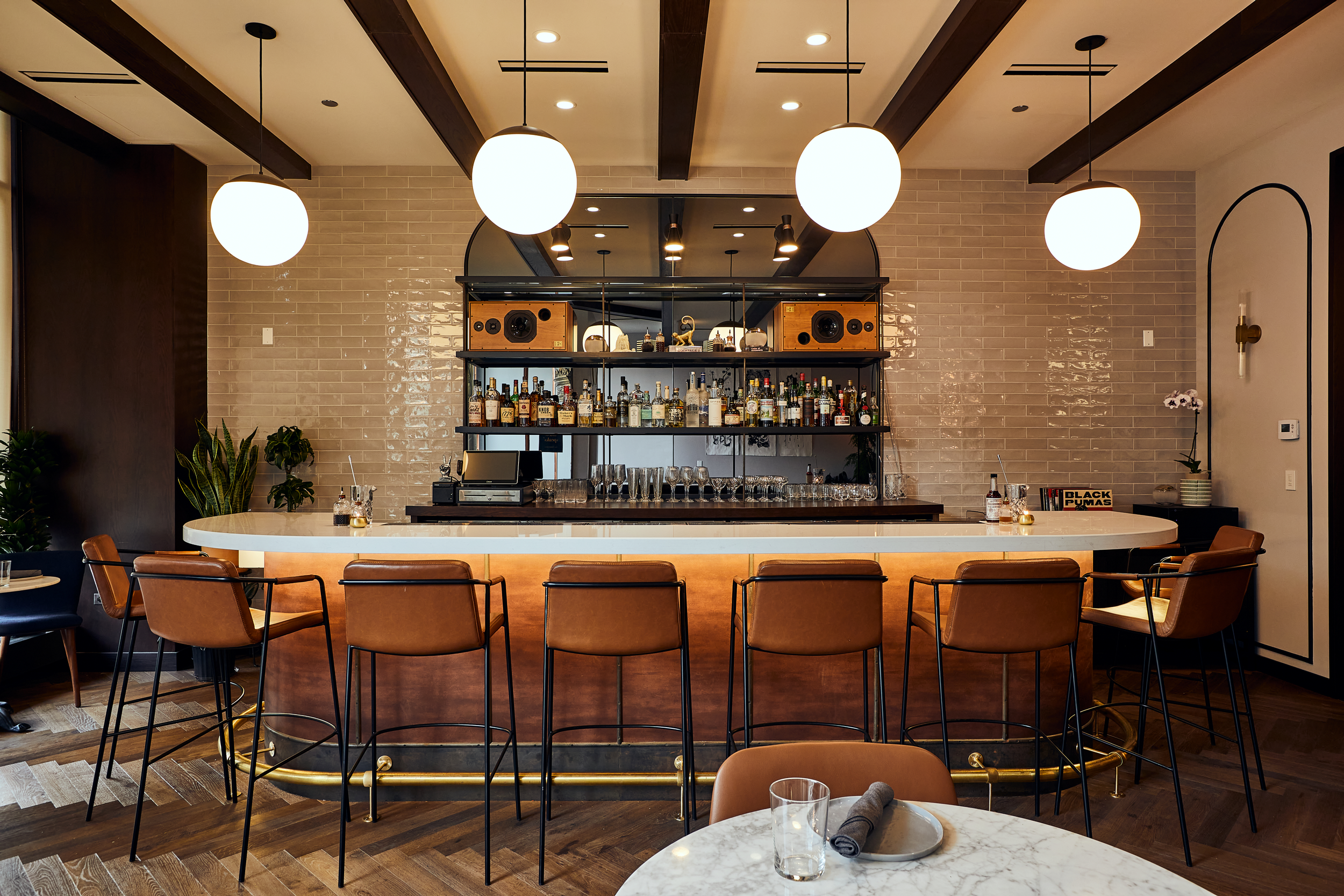 Interior of a modern bar or restaurant with a curved bar counter, chairs, a beverage shelf with liquor bottles, and decorative lighting fixtures hanging from the ceiling.