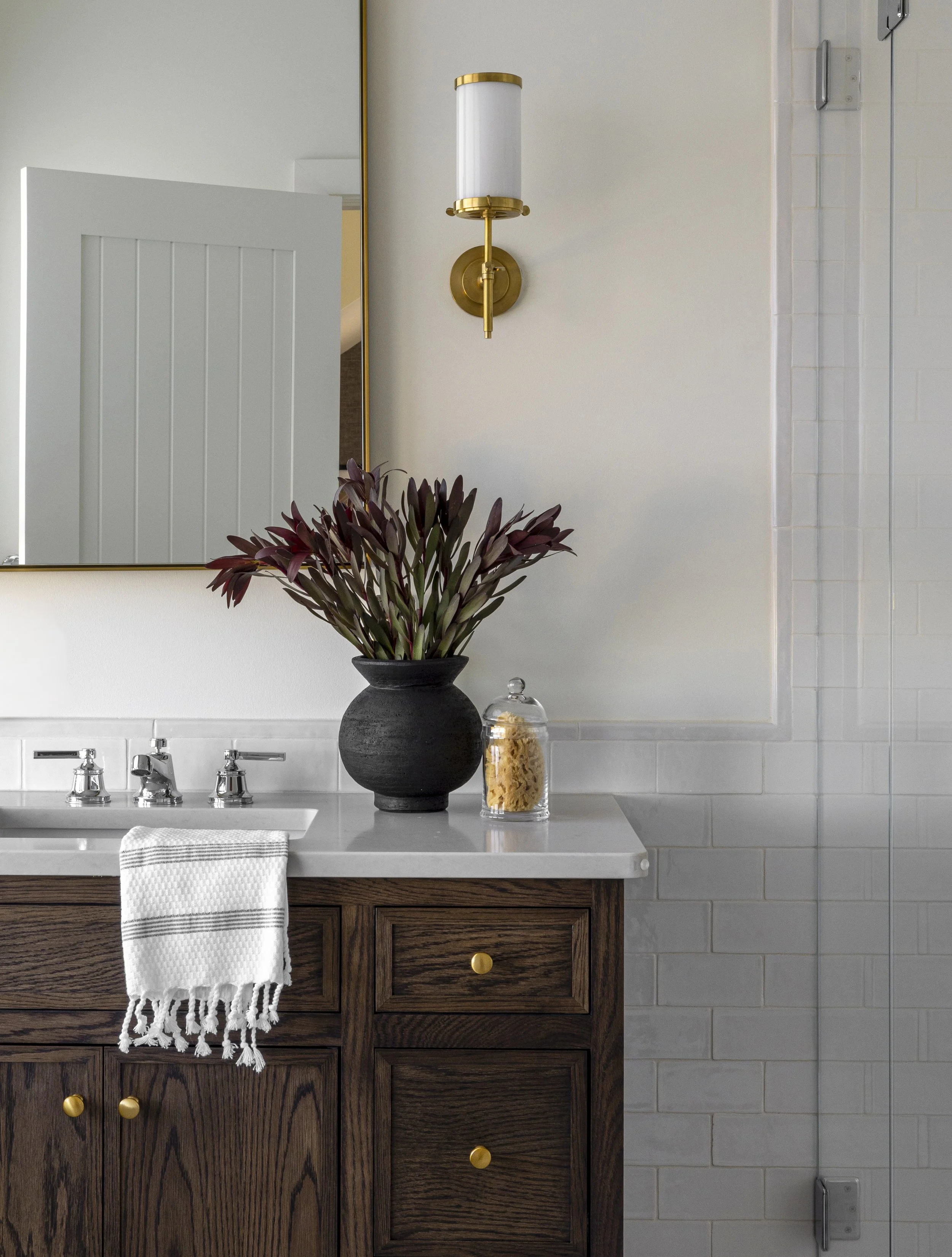 A bathroom vanity with a dark wood finish, three drawers with round gold knobs, and a white countertop with a built-in sink and silver faucets. On the counter, there is a black vase with dark purple leaves, a clear glass jar filled with pasta, and a white towel with fringe hanging from the edge of the cabinet.