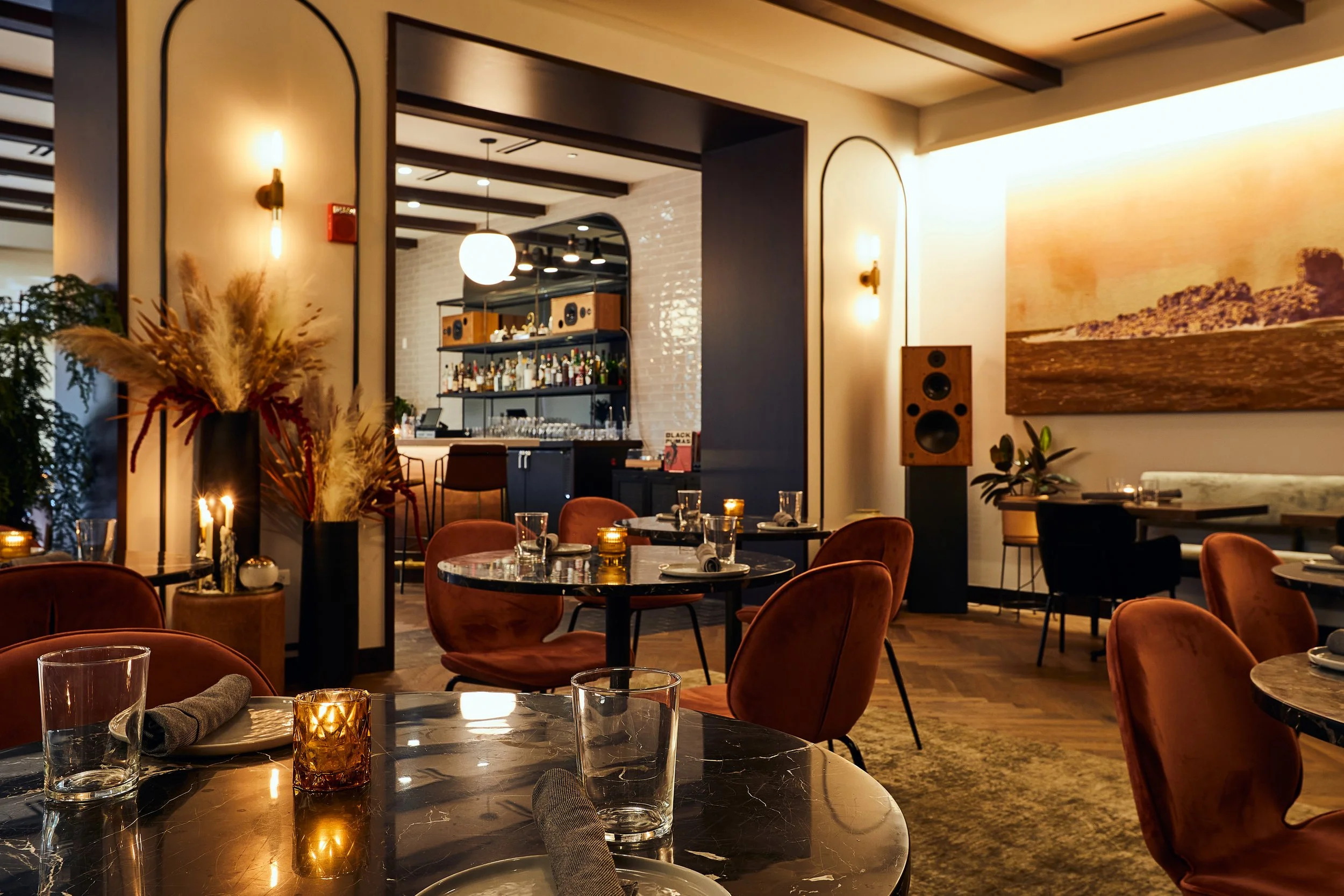 The interior of a cozy restaurant with round tables, orange chairs, candles, glasses, and napkins. There is a large painting on the wall, a speaker, and pendant lighting above a bar area in the background.