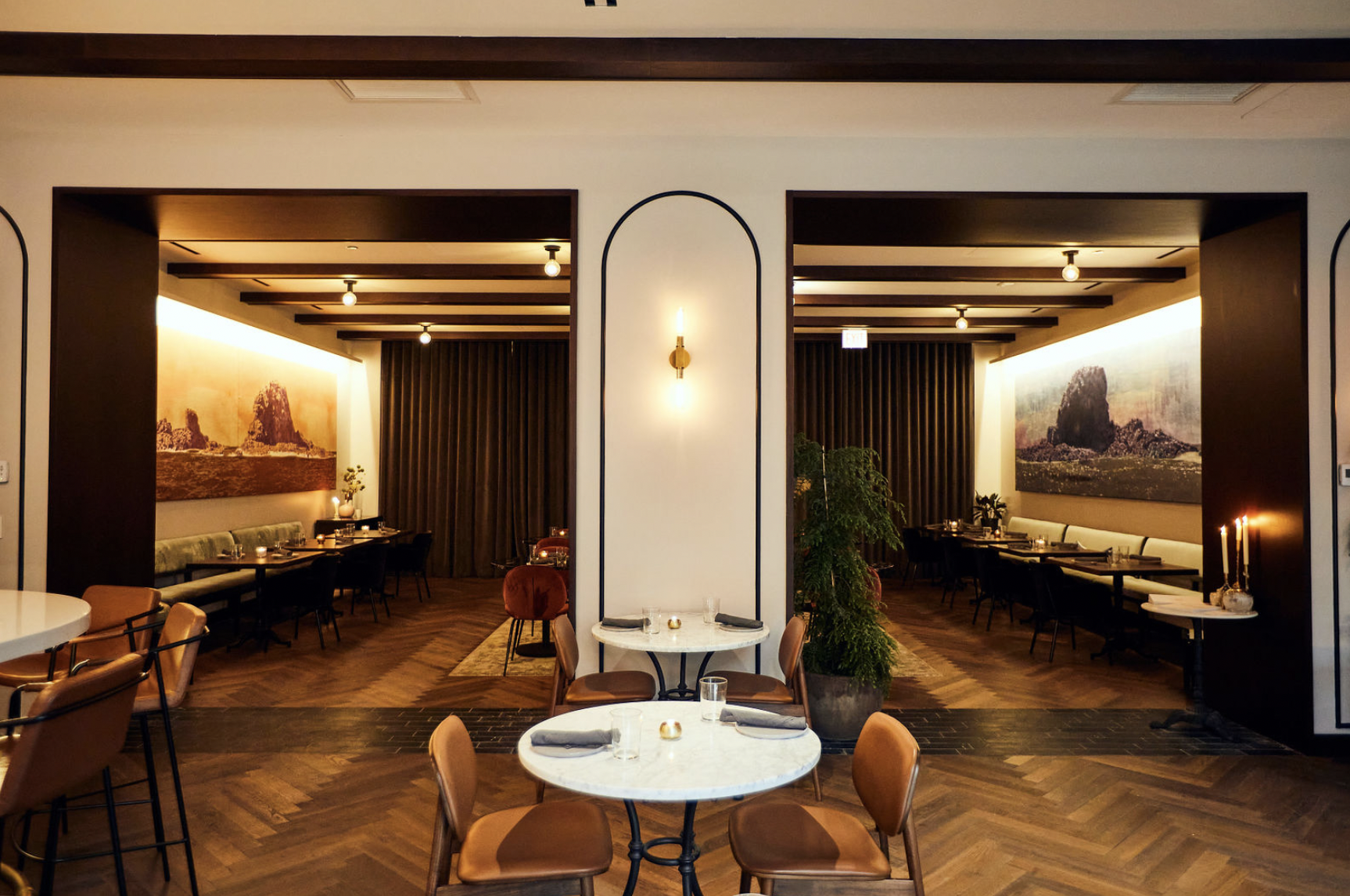 Interior of a restaurant with round tables and chairs, large landscape artwork on the walls, and a cozy lighting ambiance.