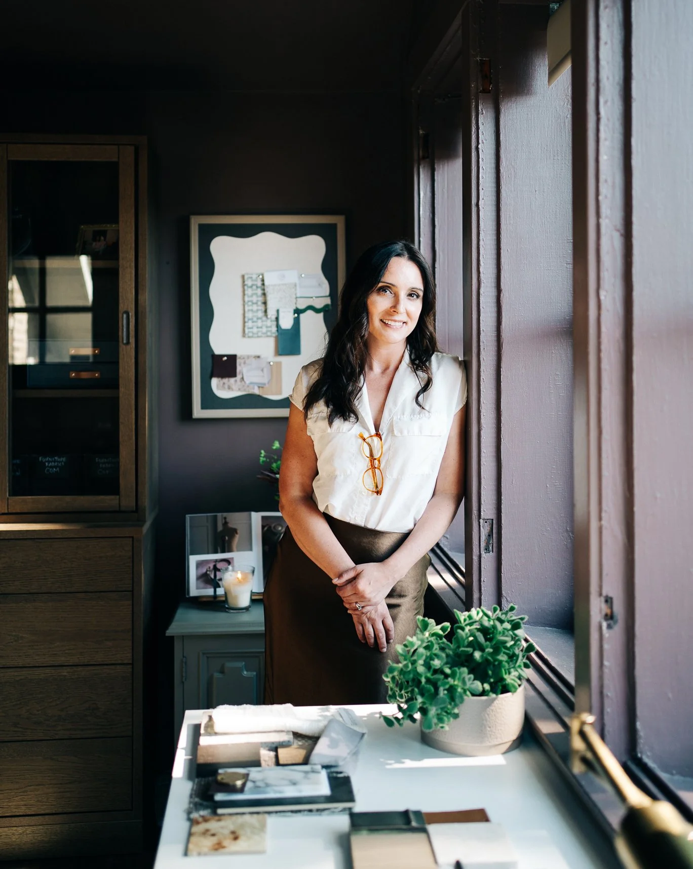 A woman with dark wavy hair smiling and standing by a window in a room filled with natural light, decorated with plants, candles, and framed photos.