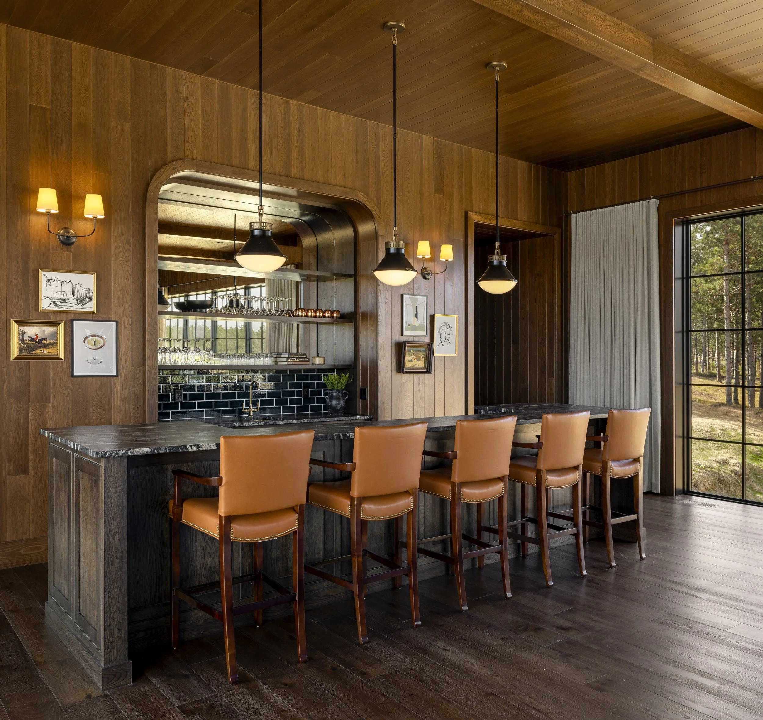 Interior view of a room with wooden walls and ceiling, pendant and wall lights, framed artwork, a window with vertical blinds, and a bar area with barstools.