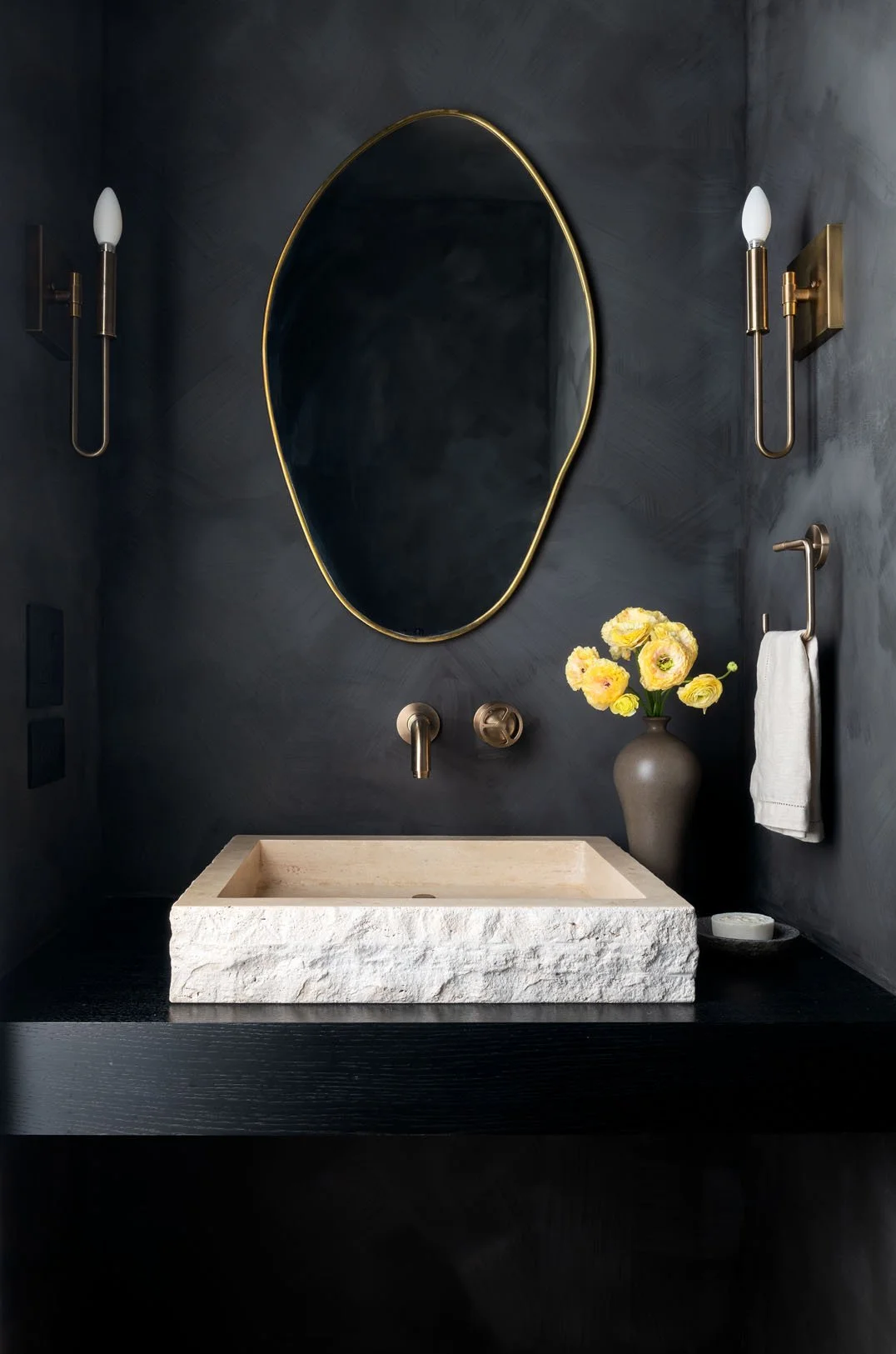 Modern bathroom with dark gray walls, an oval mirror with a gold frame, two wall sconces, a beige stone sink, a vase with yellow flowers, and a white towel on a hook.
