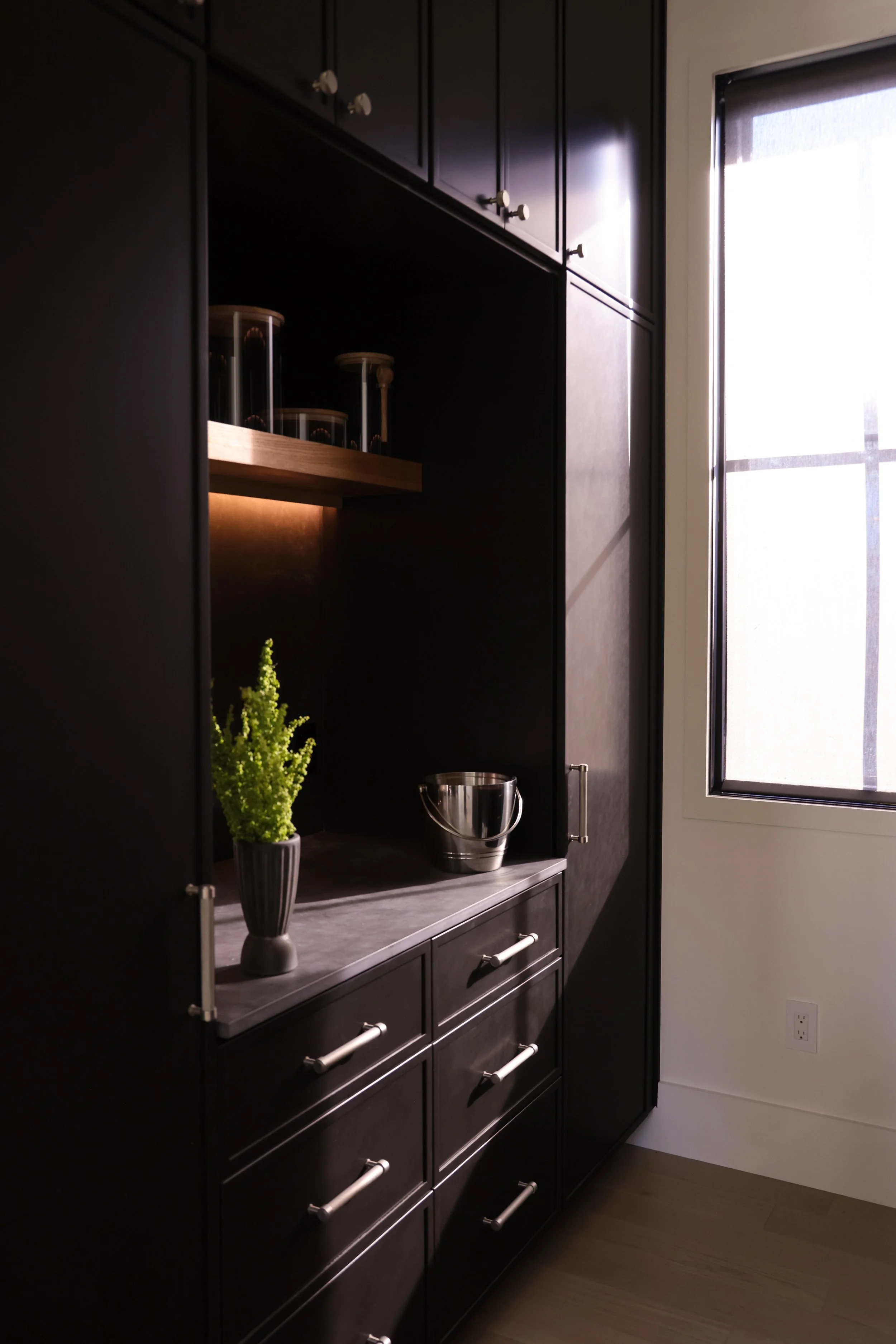 A black kitchen cabinet with silver handles, a wooden shelf with glass jars, a silver bucket, a potted green plant, near a window with light coming through.