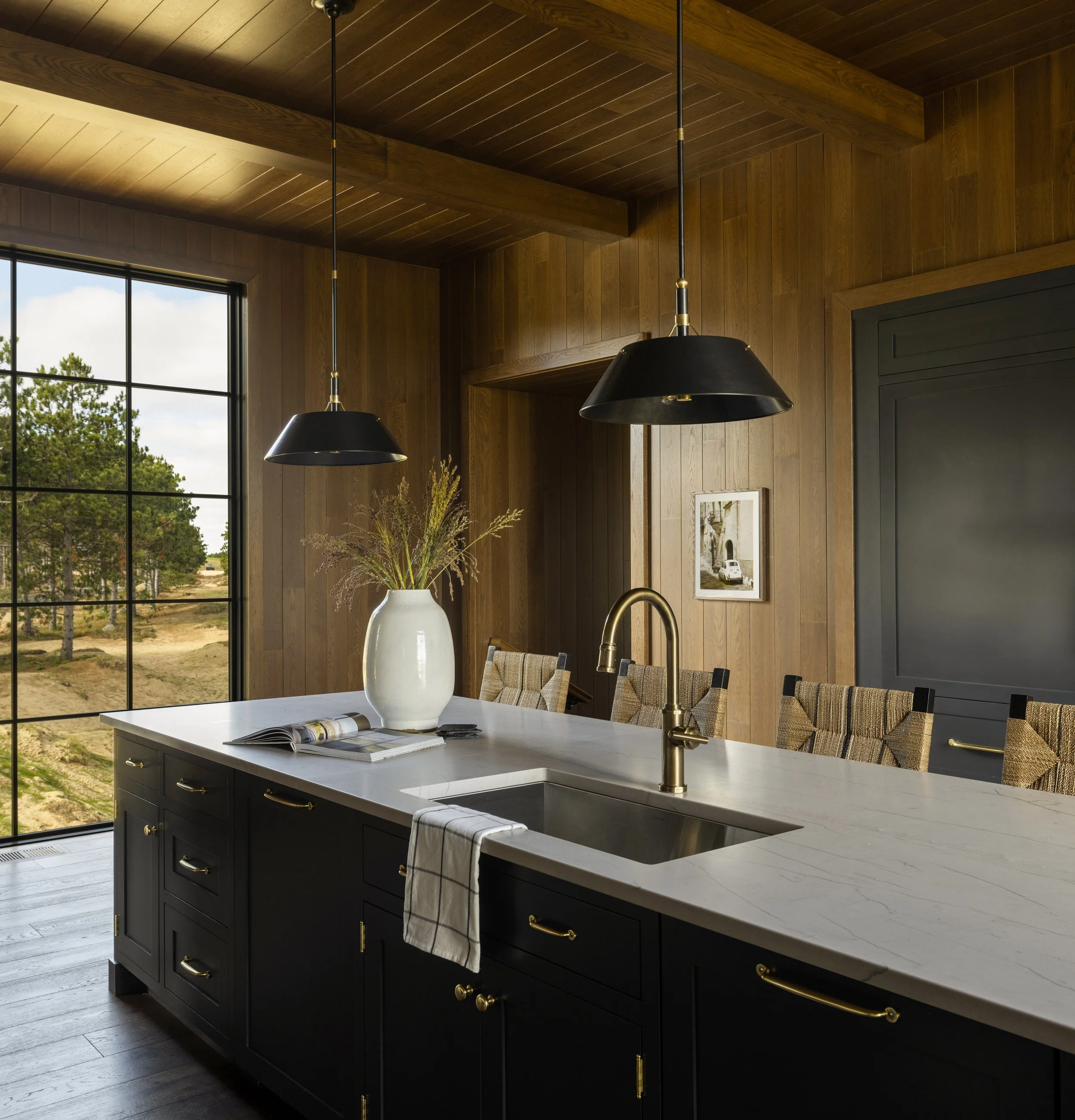Modern kitchen with a black island, gold hardware, marble countertop, large window with a view of trees and a construction site, wooden walls, and pendant lights.