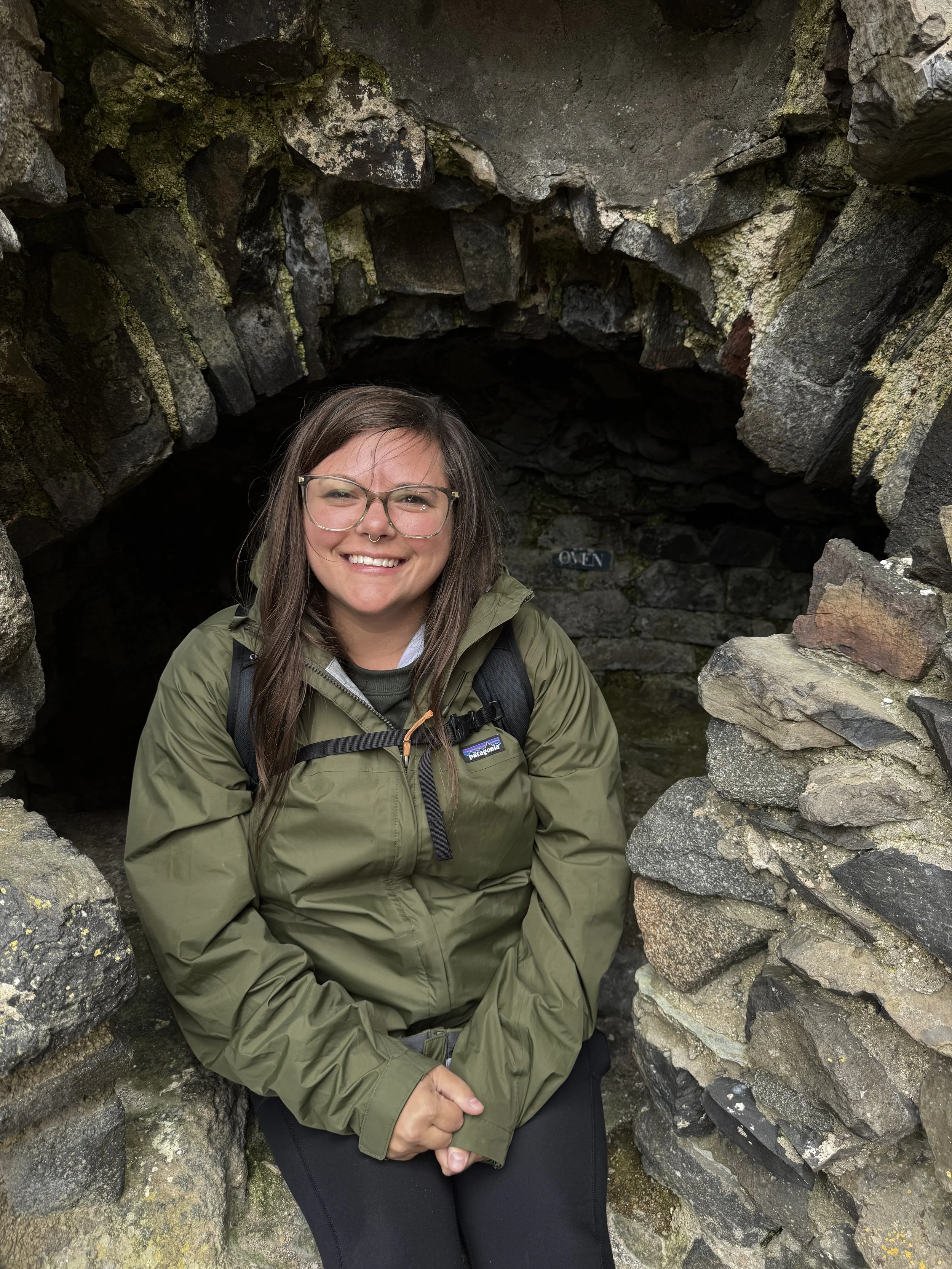 A smiling woman with glasses and a green jacket sitting inside a small stone cave, surrounded by rocks.
