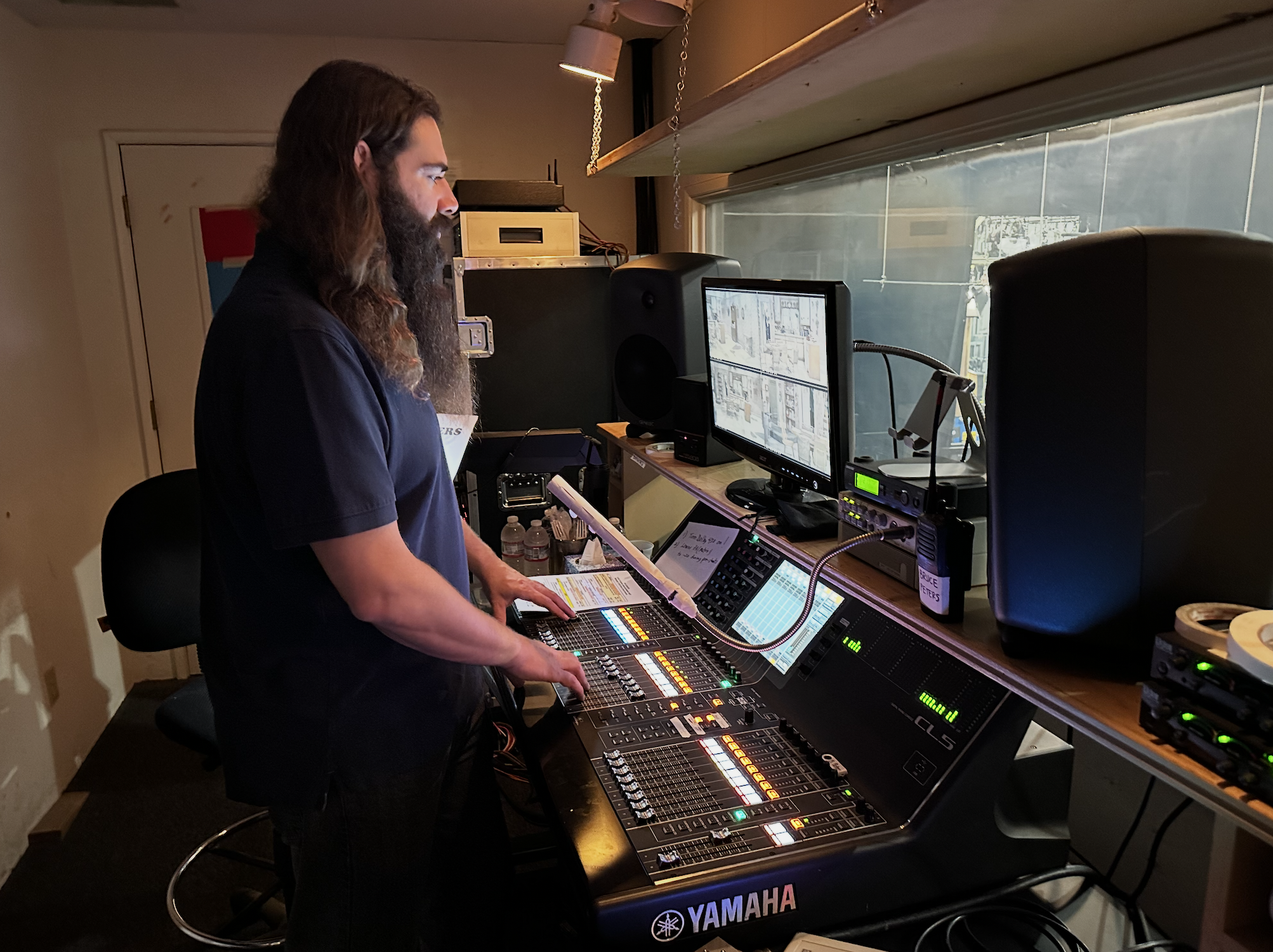 A man with long hair and a beard operating sound mixing equipment in a studio, with a monitor, speakers, and a large window in the background.