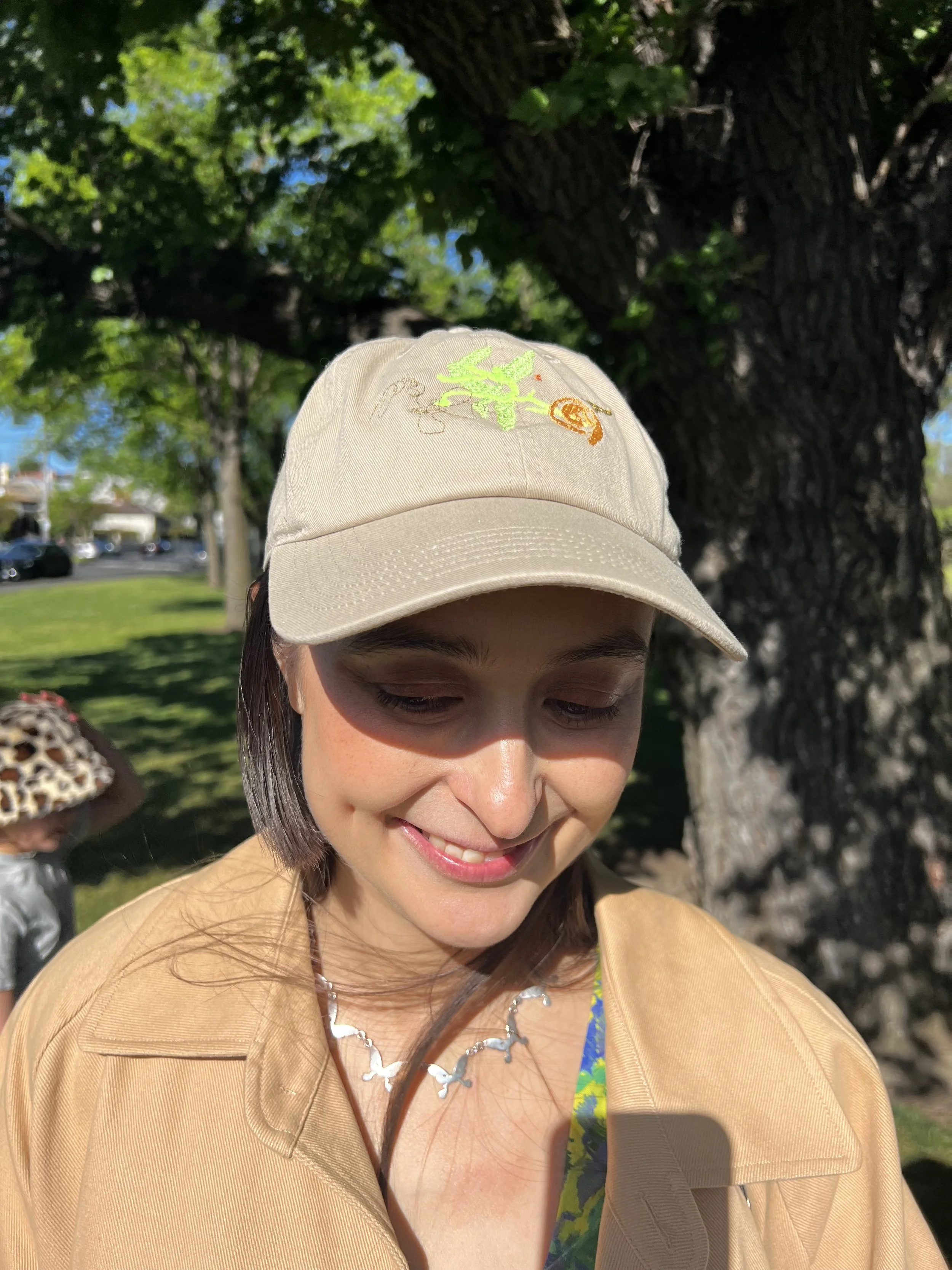 A woman wearing a beige hat with embroidered green and orange details, smiling while standing outdoors near a tree with green foliage.