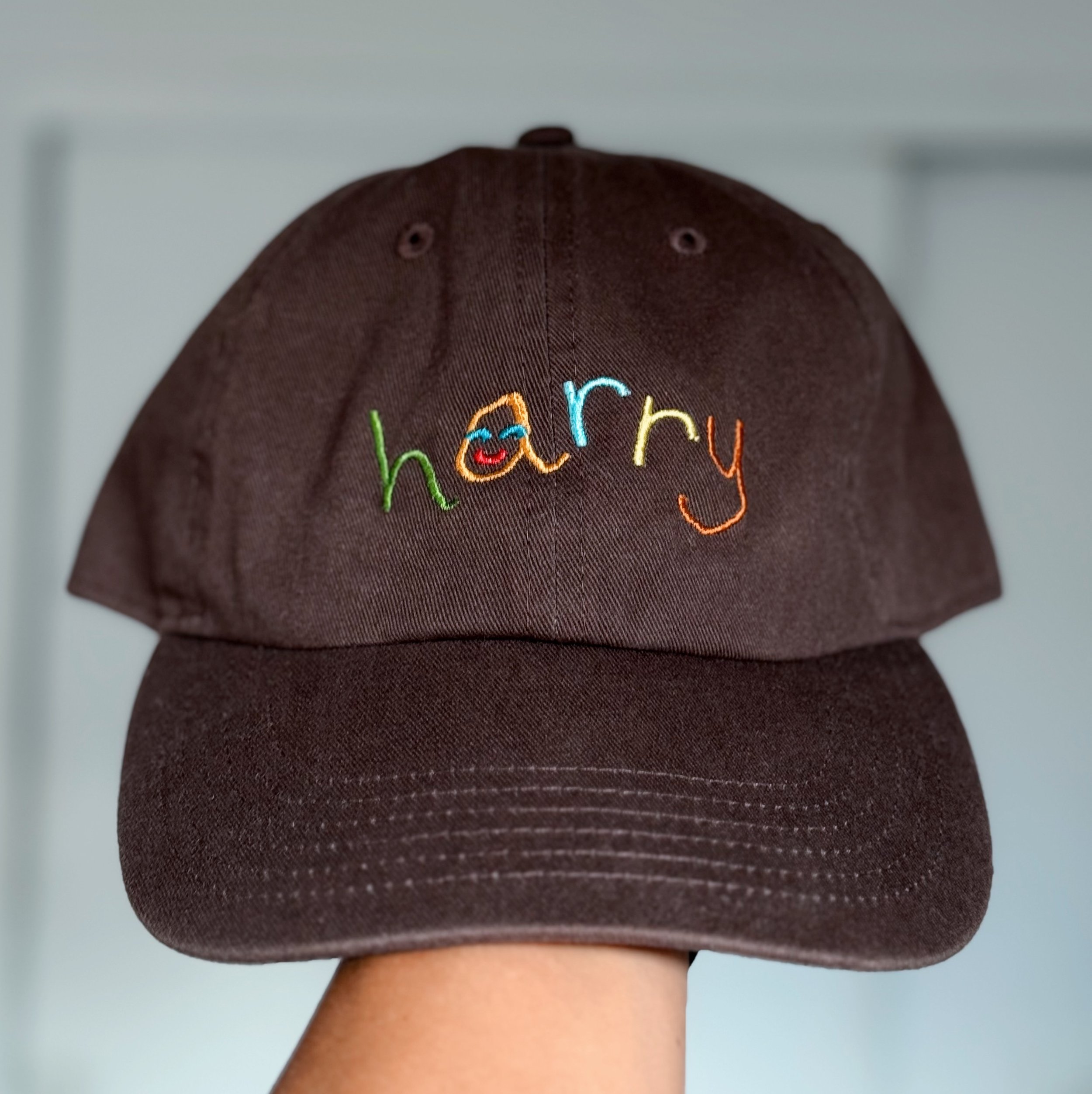 Close-up of a dark brown baseball cap with colorful embroidered text reading 'happy' and a smiling face, worn backwards.