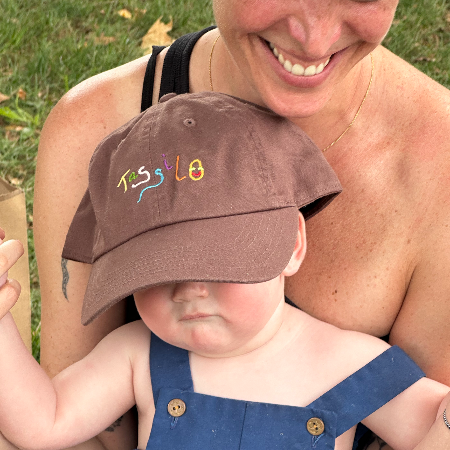 Close-up of an adult woman smiling, holding a young child. The woman is wearing a black tank top and a thin gold necklace. The child is shirtless, wearing a navy blue outfit with gold buttons and a brown hat with colorful embroidery that says "TassLo