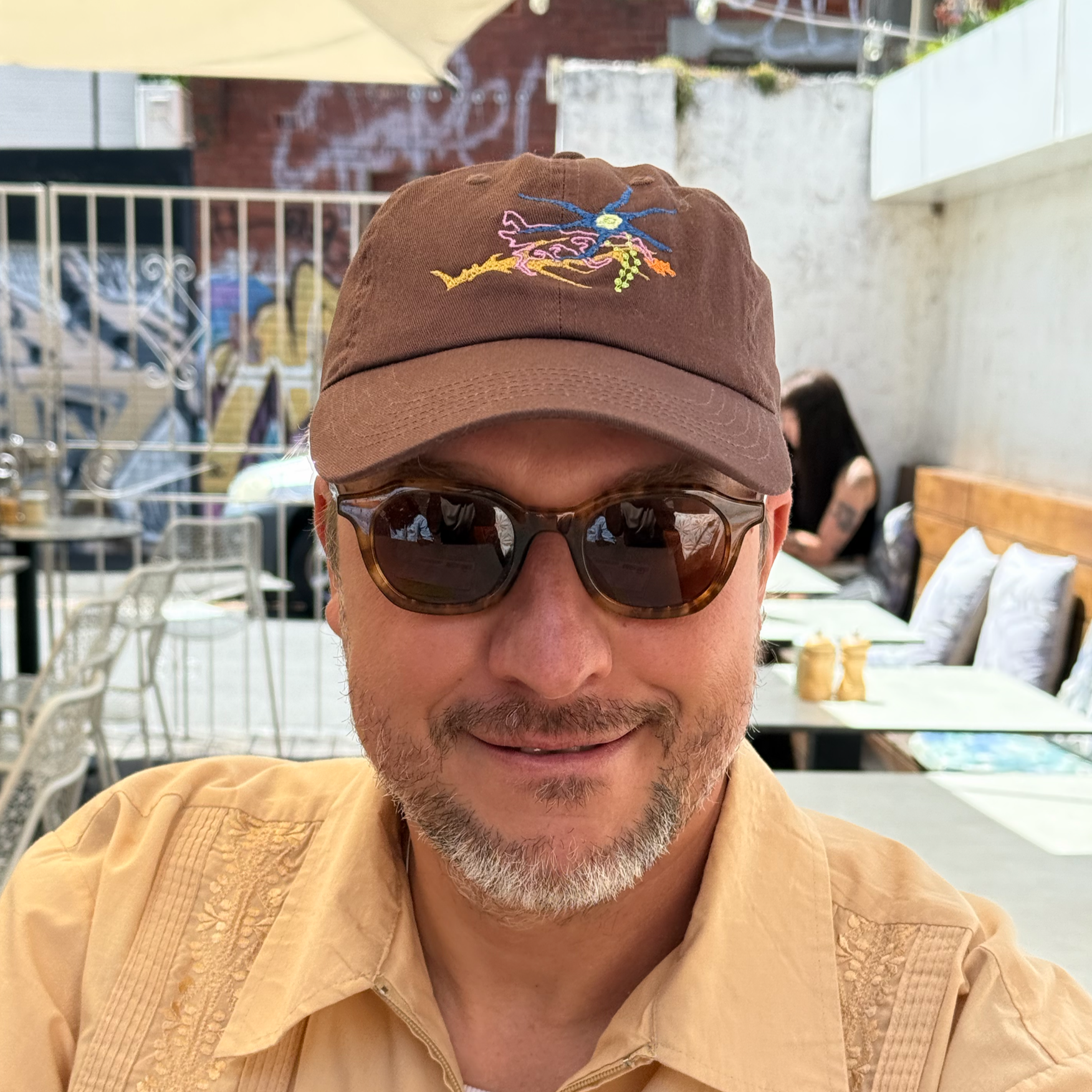 Close-up of a man wearing sunglasses, a brown cap with a colorful embroidered design, and a light yellow shirt, smiling at an outdoor cafe with tables, chairs, and a woman in the background.
