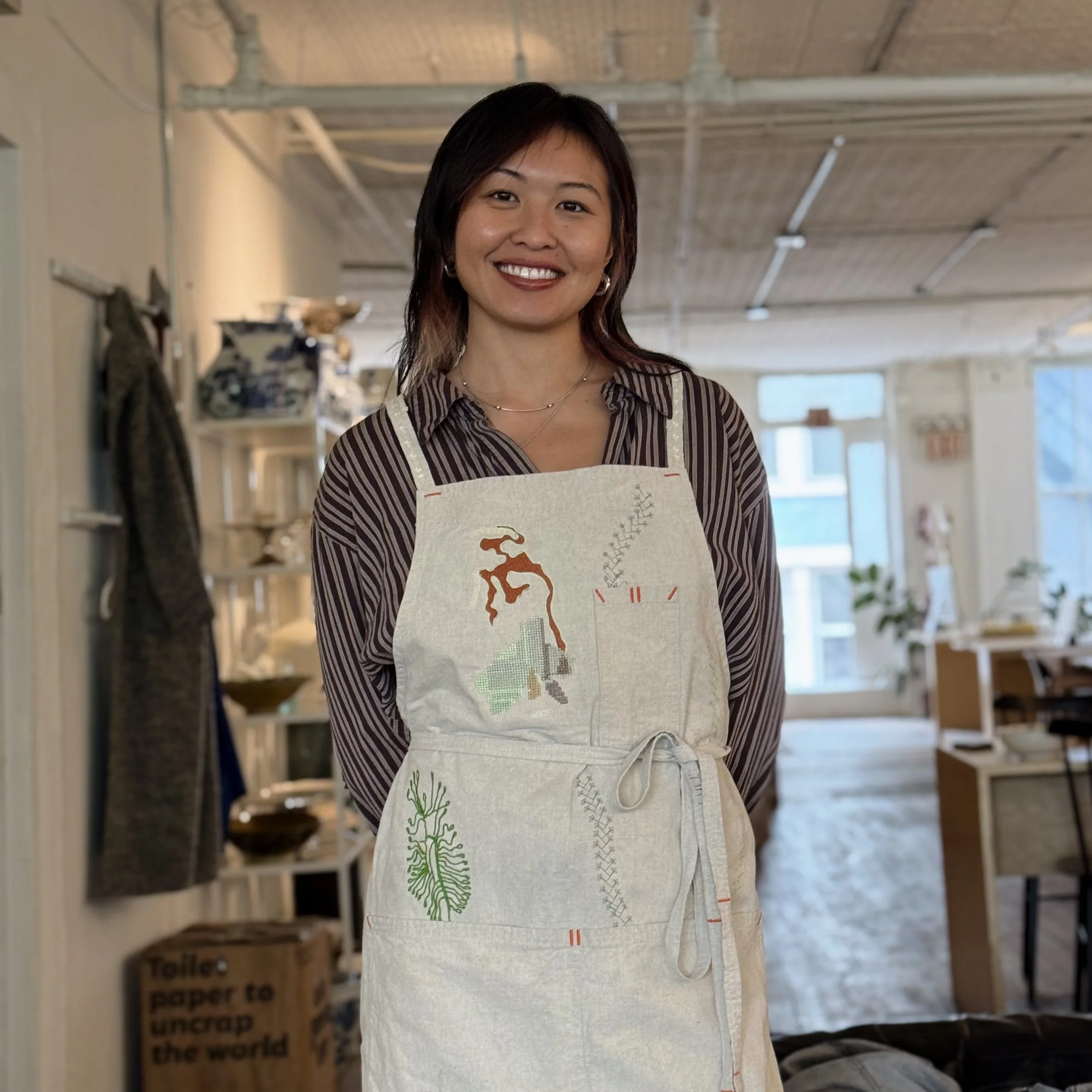 A smiling woman with dark hair wearing a striped shirt and an apron, standing indoors with shelves and boxes in the background.