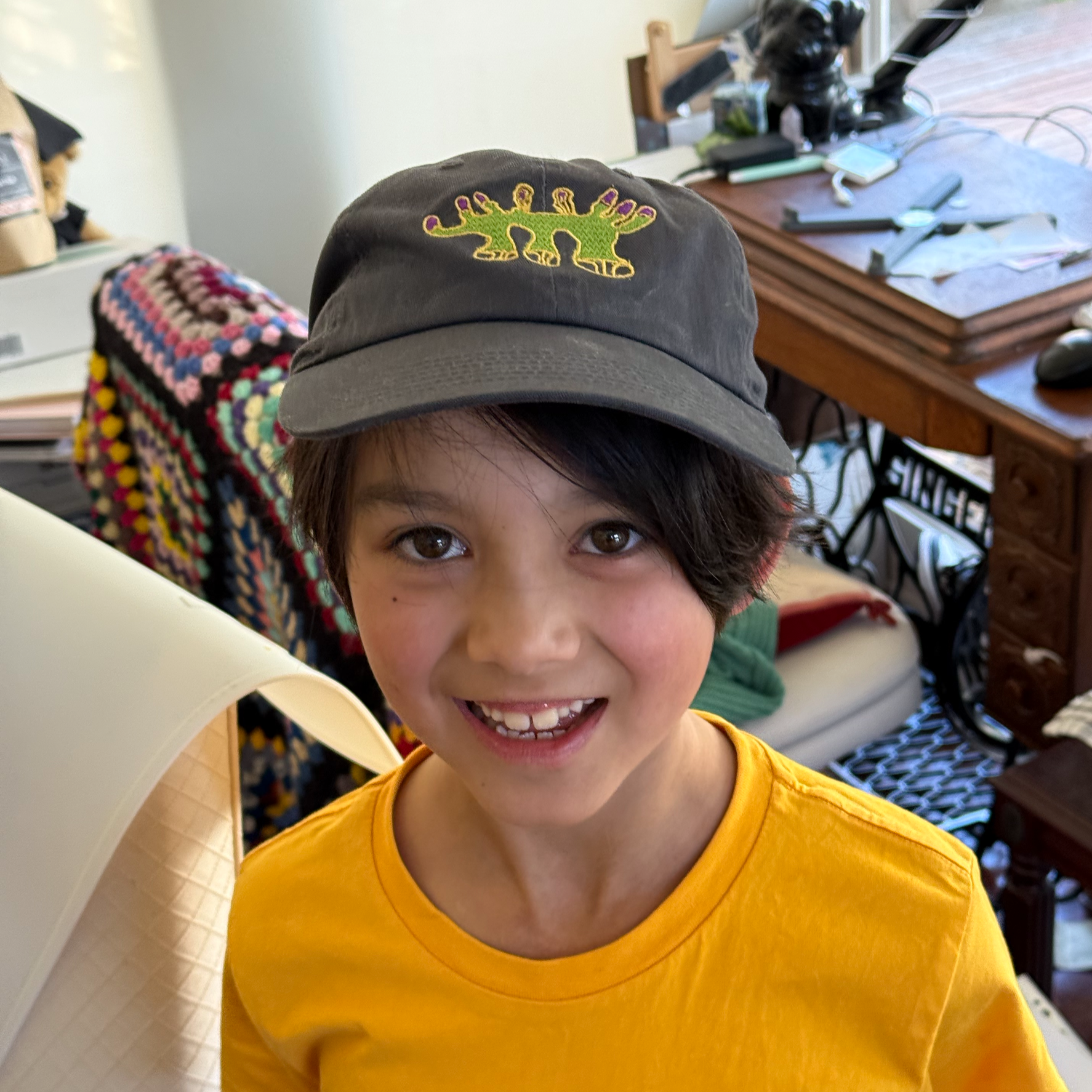 A child wearing a yellow T-shirt and a gray baseball cap with a dinosaur embroidery, smiling and showing teeth, indoors with furniture and personal items in the background.