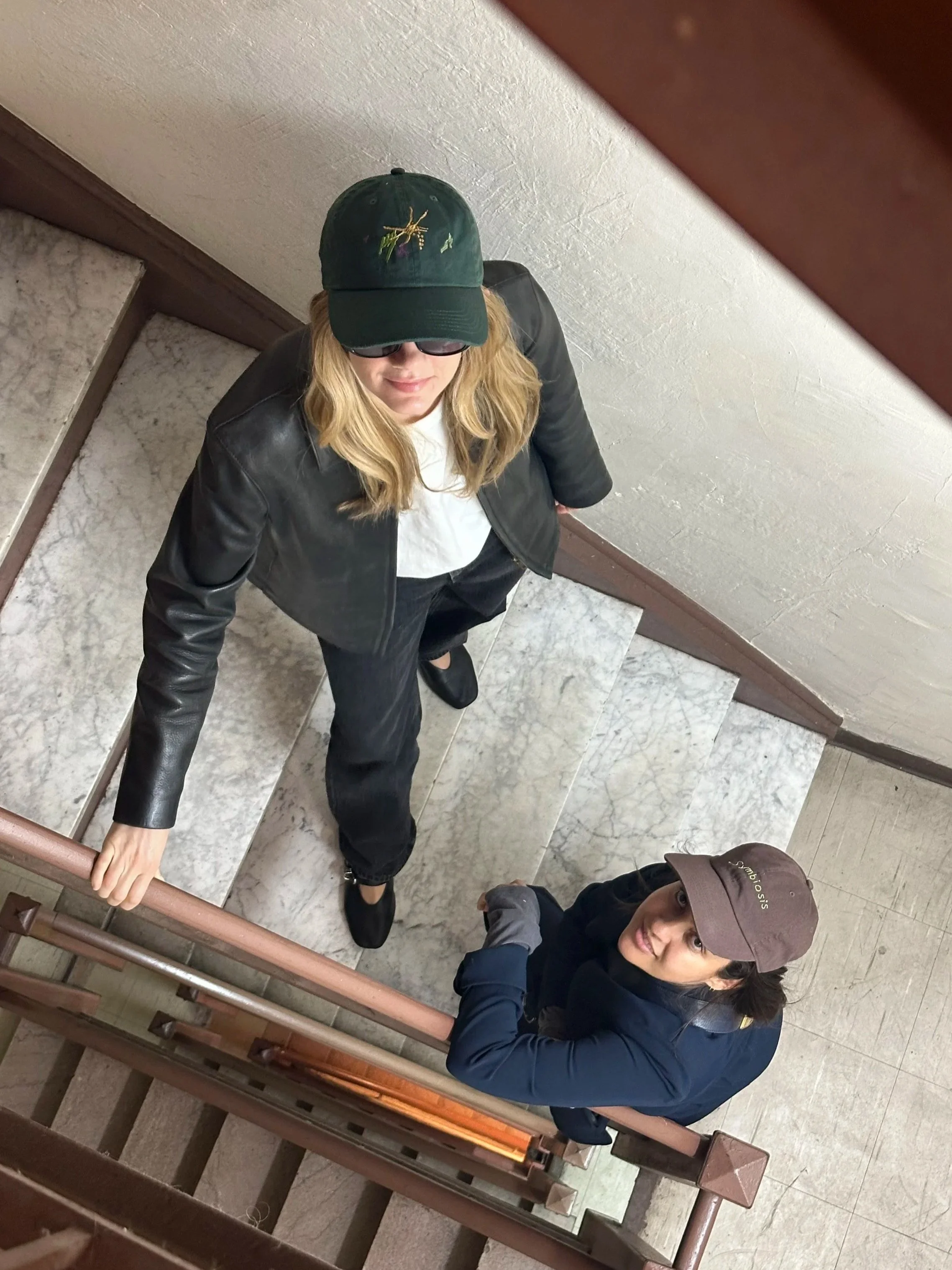 Two women are looking up at the camera at the top of a staircase. One woman, with long blonde hair, is wearing a black leather jacket, white shirt, black pants, and a green baseball cap with colorful embroidery. The other woman, with dark hair, is we