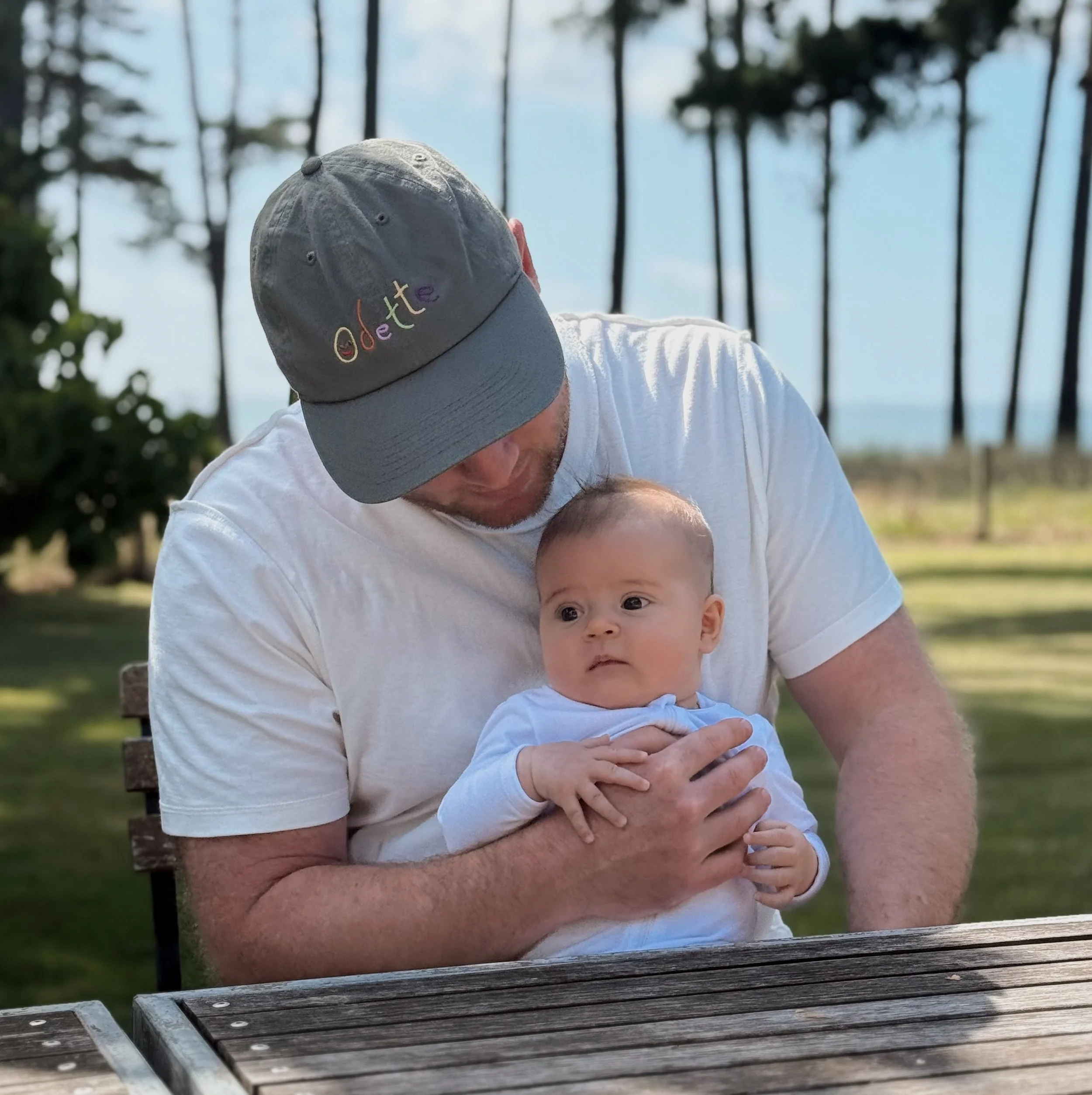 A man in a gray cap embroidered with the word 'dedette' and a white T-shirt holds a baby on his lap on a park bench, with palm trees in the background.