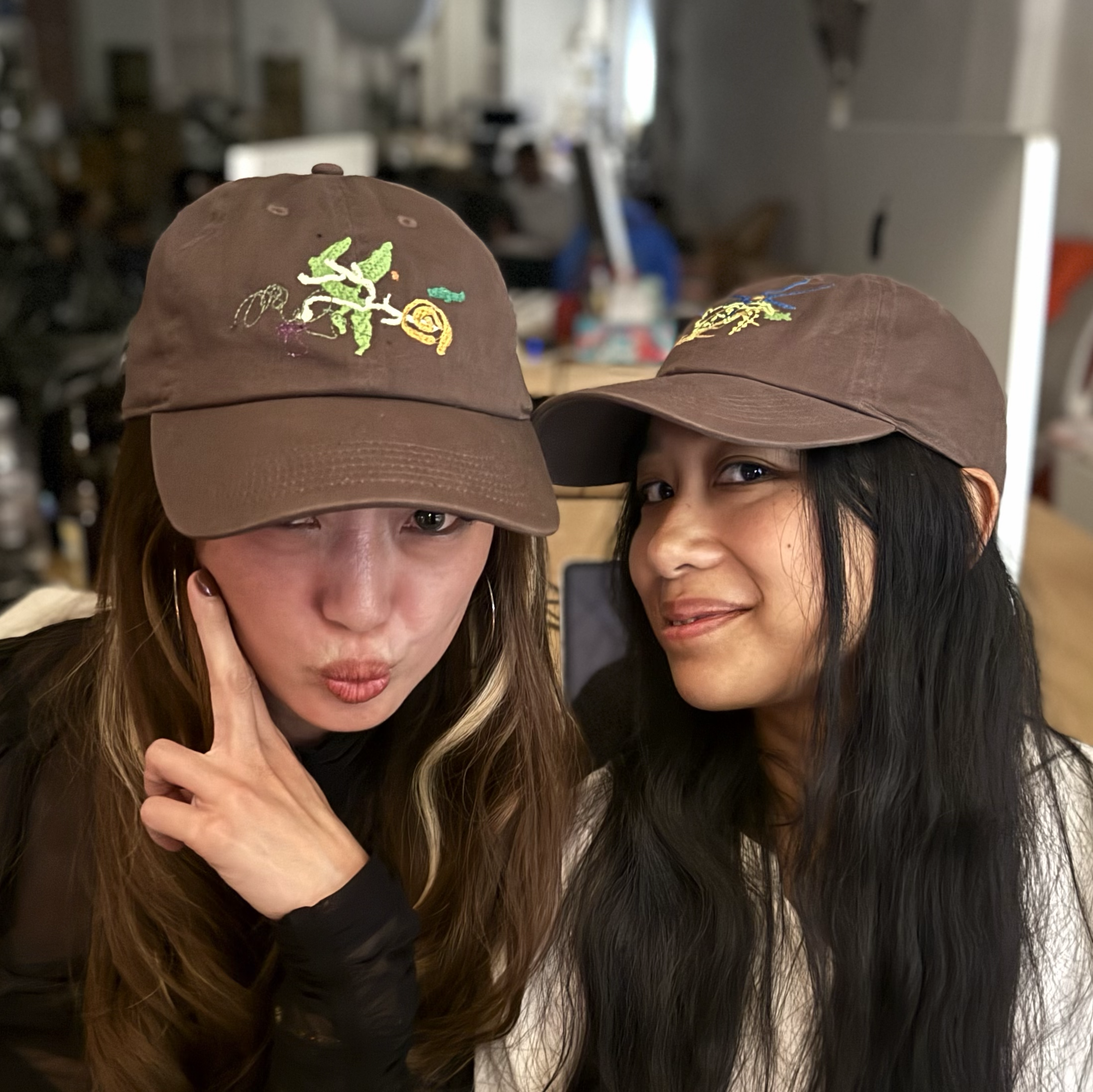 Two women wearing brown hats with colorful embroidery, smiling and close together indoors.