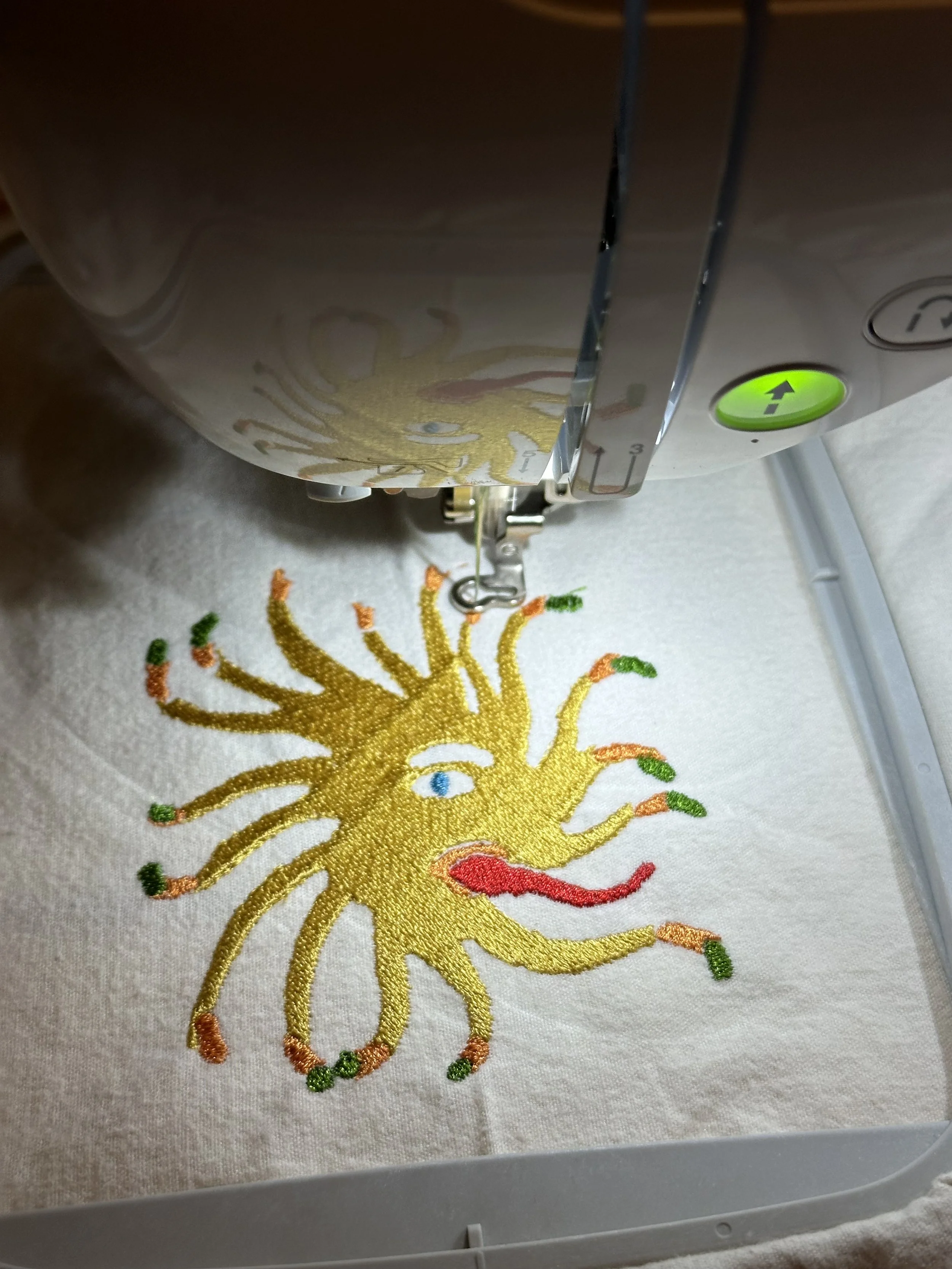 A sewing machine stitching a colorful embroidered design of a sun with a red mouth, yellow rays, green and orange tips, and a single eye on a light-colored fabric.