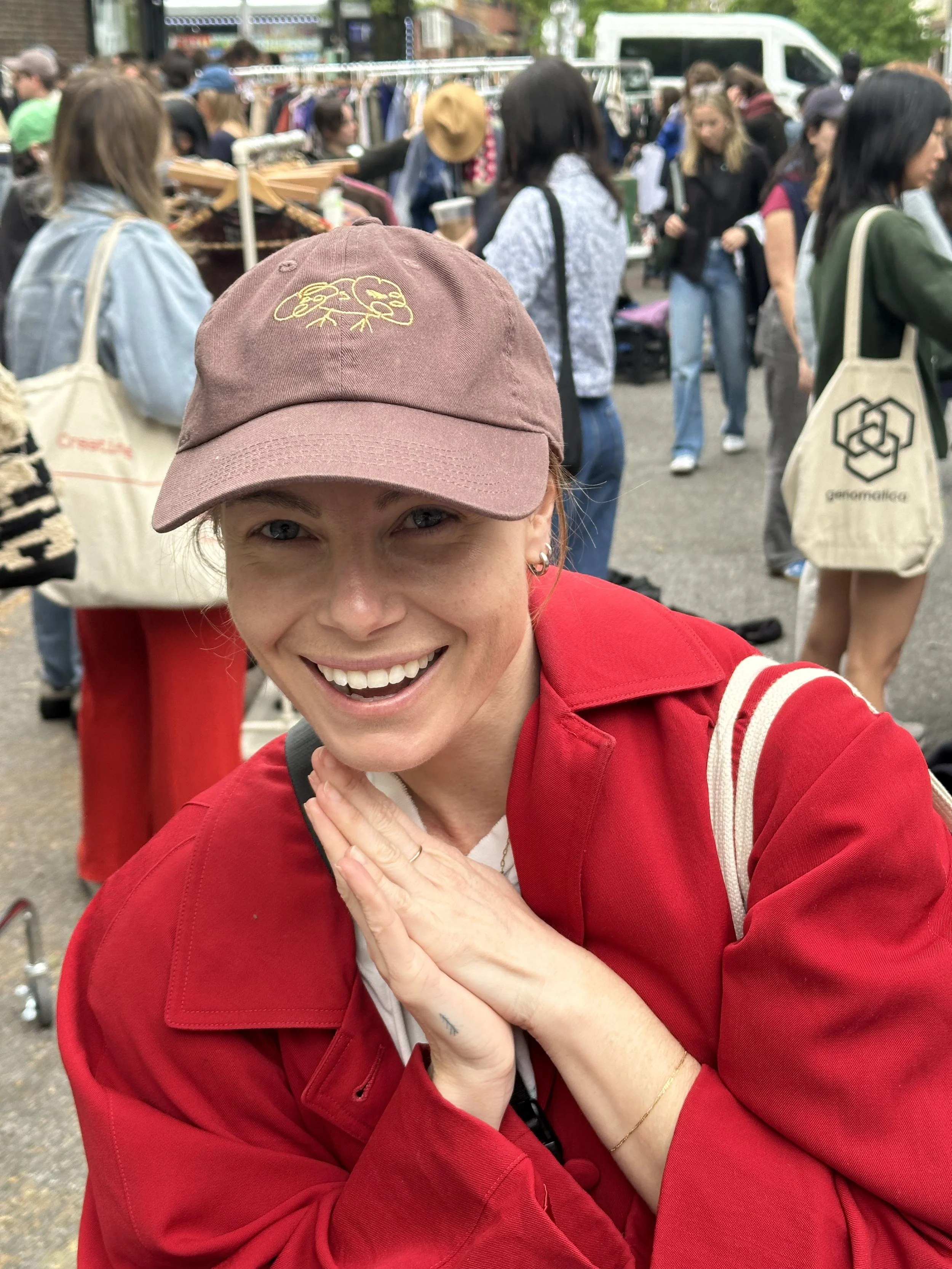 A smiling woman in a red jacket and pink baseball cap taking a selfie at an outdoor market with people browsing stalls in the background.