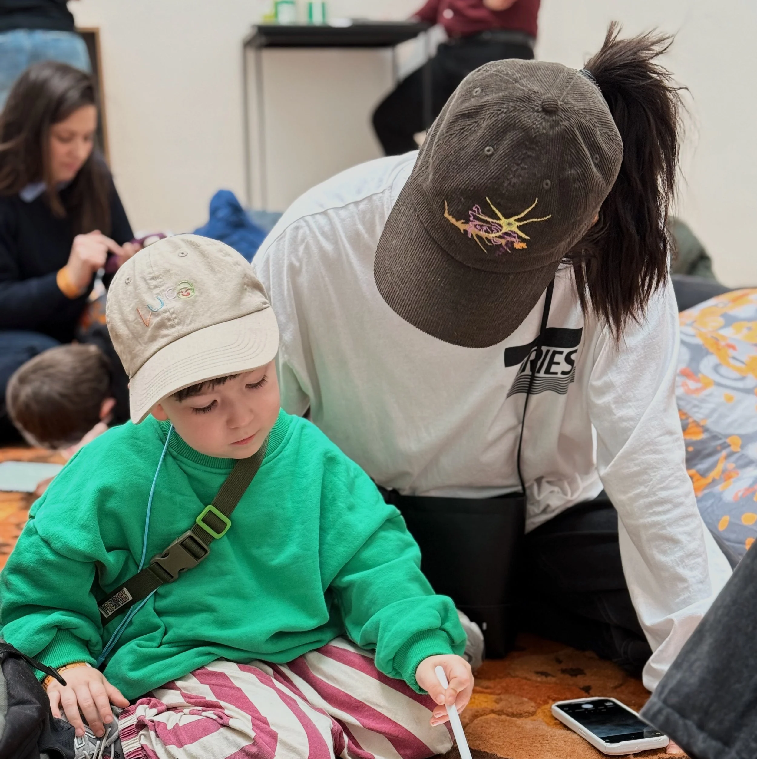 Children sitting on the floor, one in a bright green sweatshirt and beige cap, and others visible in the background, engaged with mobile phones in an indoor setting.