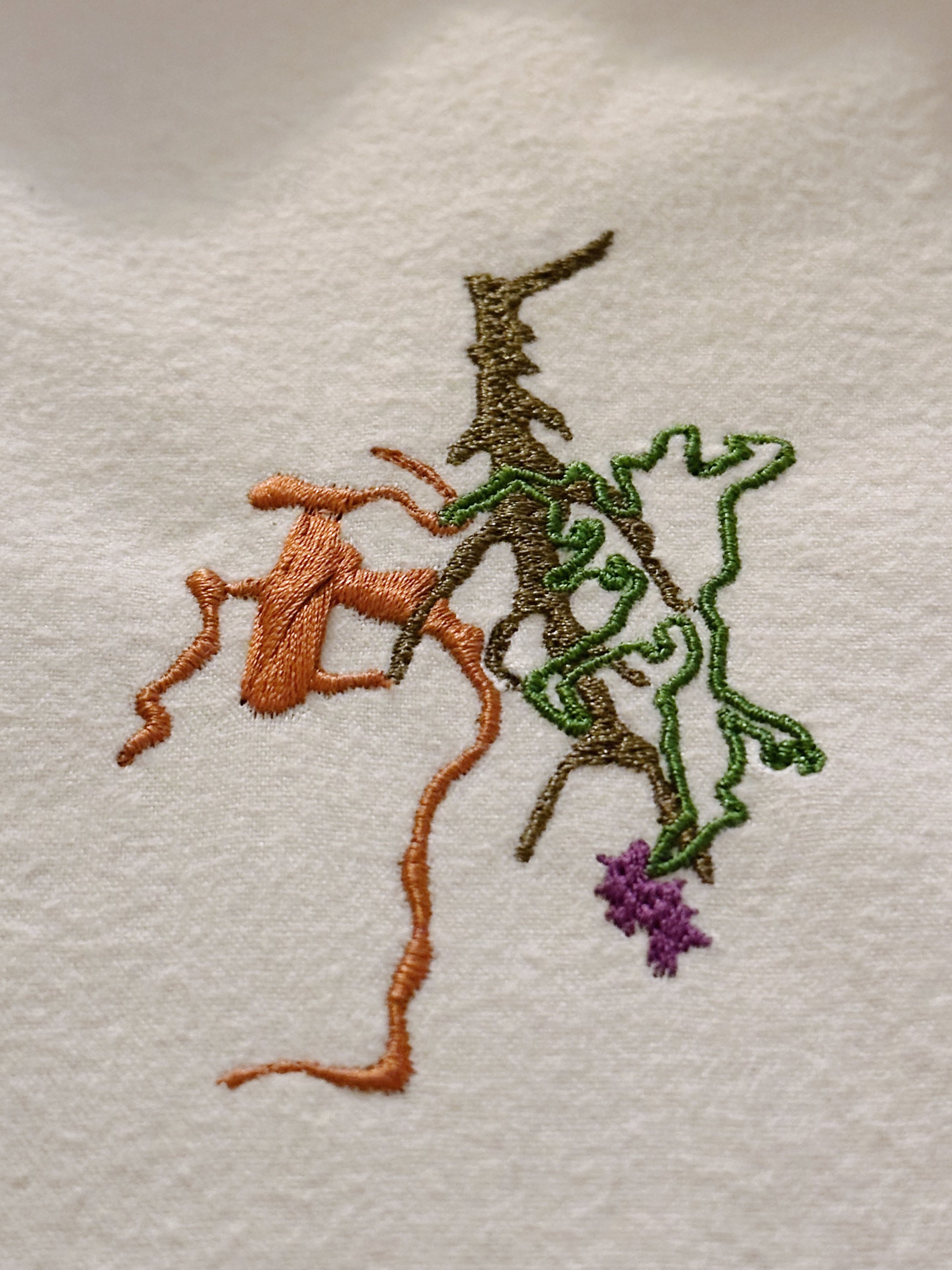 Close-up of embroidered outline of two overlapping human profiles on fabric, one in black and the other in green, with a purple flower embroidery at the bottom.