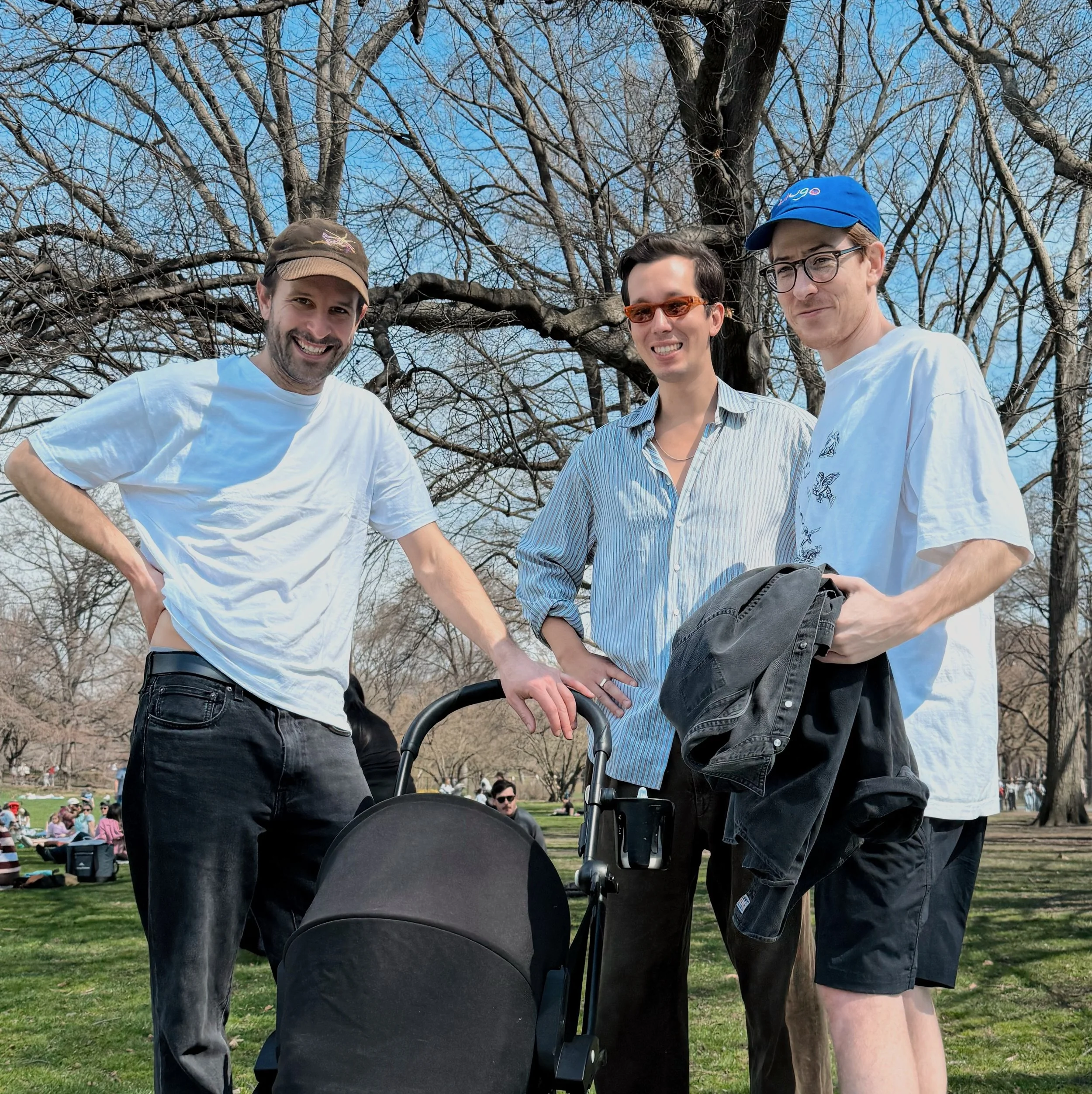 Three men standing outdoors in front of a leafless tree, smiling at the camera, with a crowd sitting on the grass in the background. The man on the left is wearing a brown cap and a white t-shirt, the man in the middle is wearing orange sunglasses an