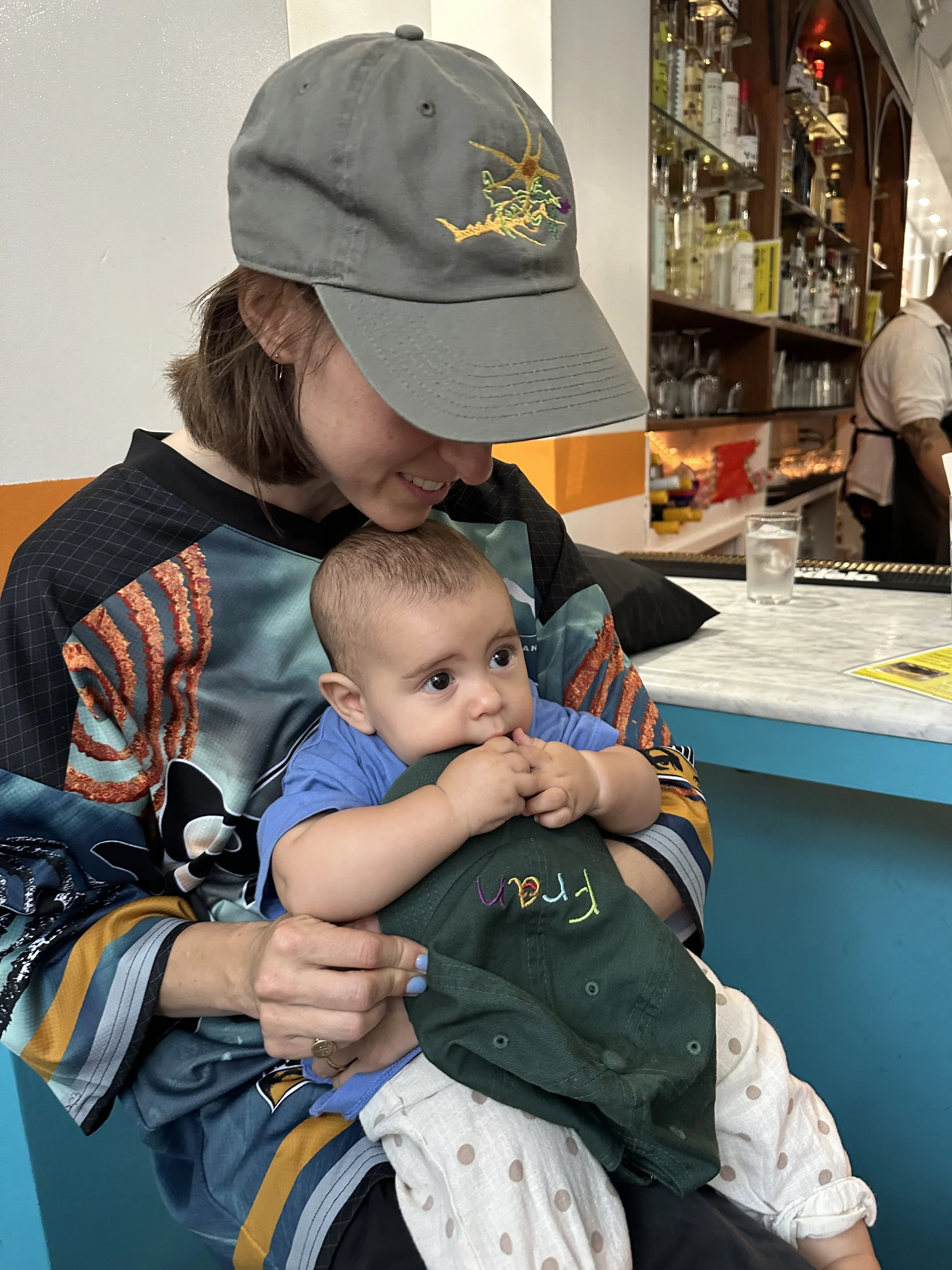 A woman wearing a gray cap with embroidery of a bird and colorful plants, smiling as she holds a young child with big eyes, wearing a blue shirt and polka-dot white pants.