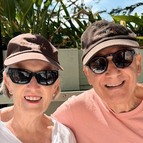 An elderly woman and man smiling outdoors, both wearing sunglasses and hats, with green foliage in the background.