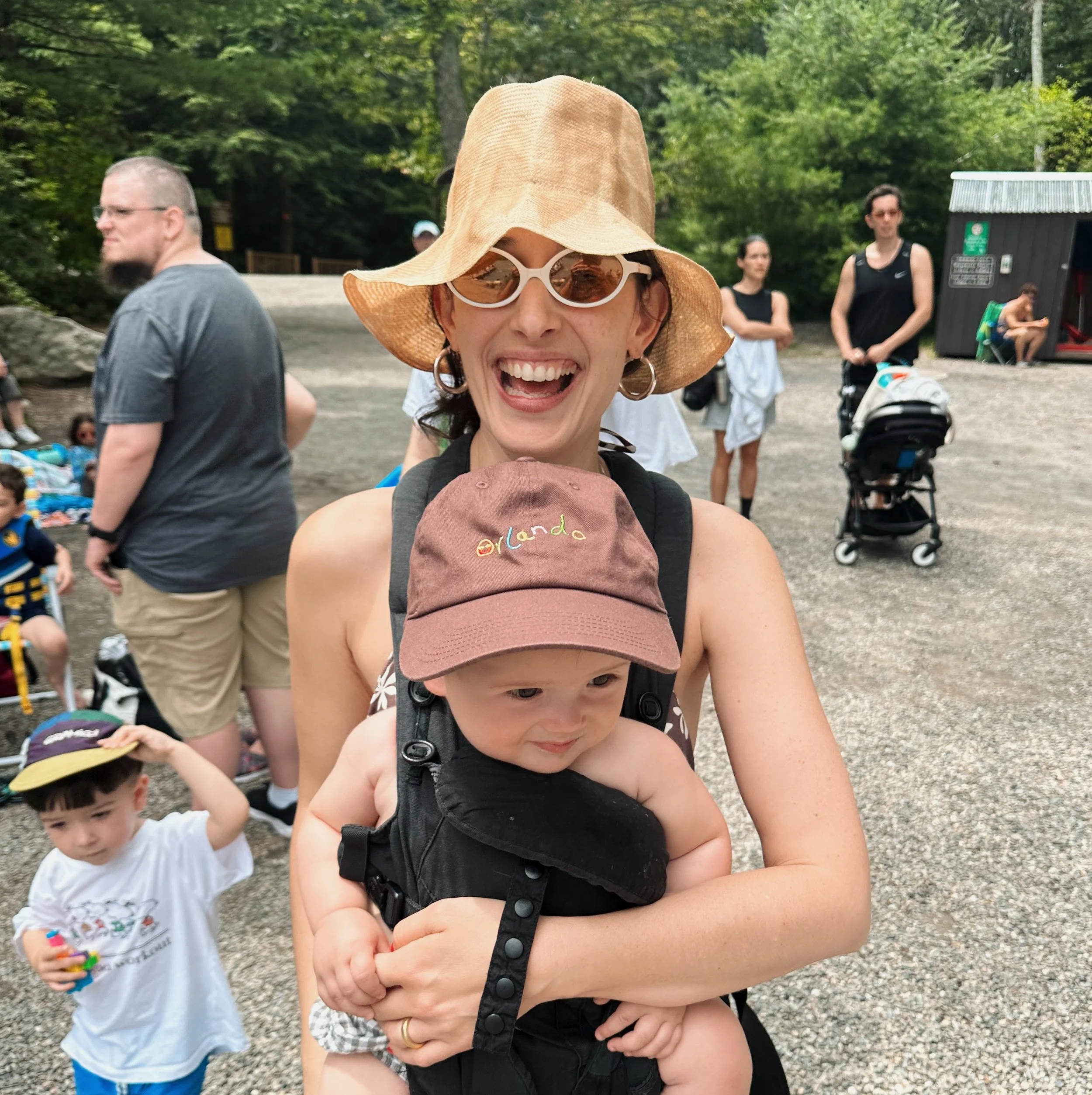 A woman smiling with sunglasses and a wide-brimmed hat, holding a child with a hat in an outdoor park where other people are gathered.
